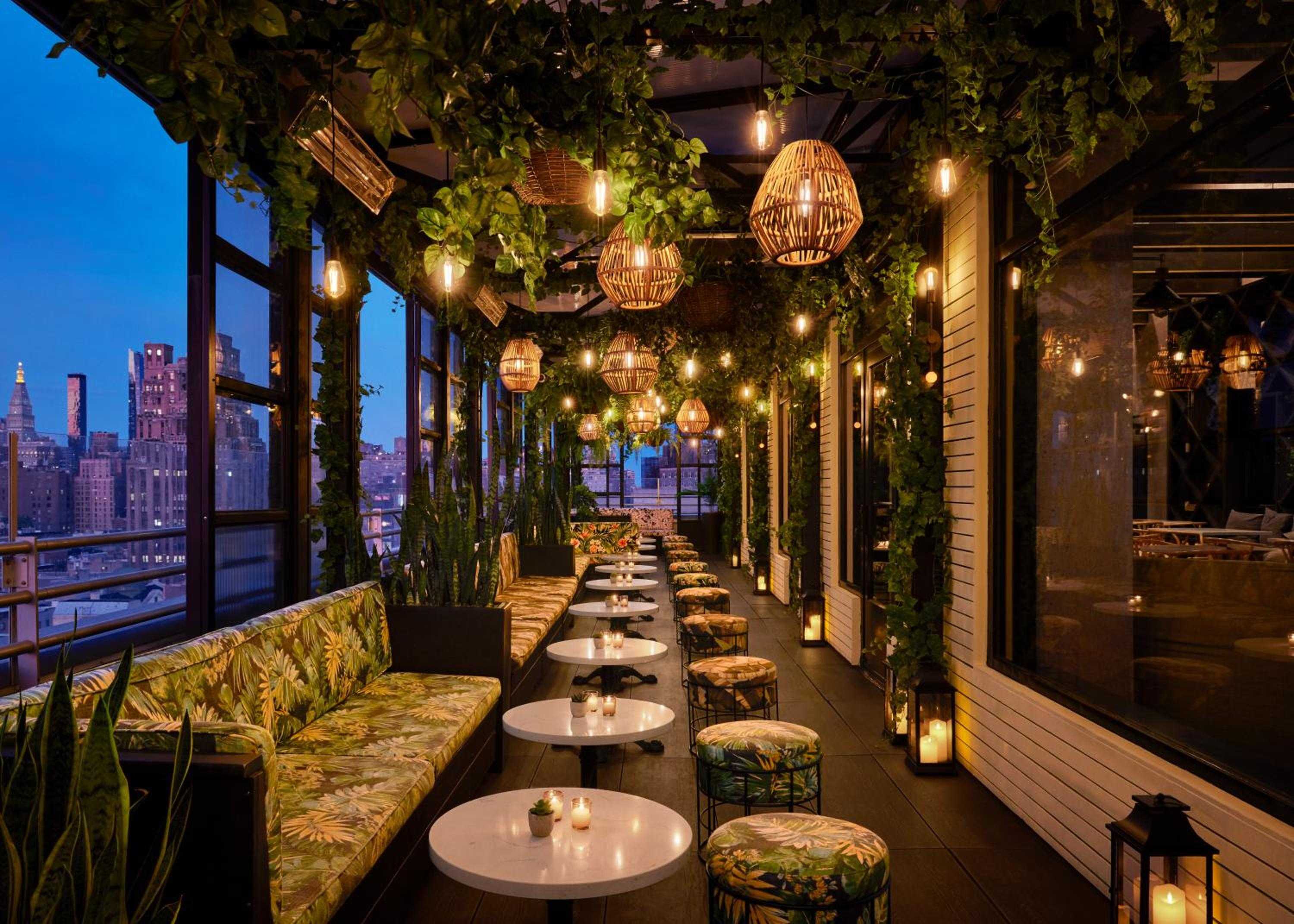 Wicker pendant lights cast golden shadows over tropical-print seating as Manhattan's skyline glitters beyond this enchanting rooftop garden retreat.