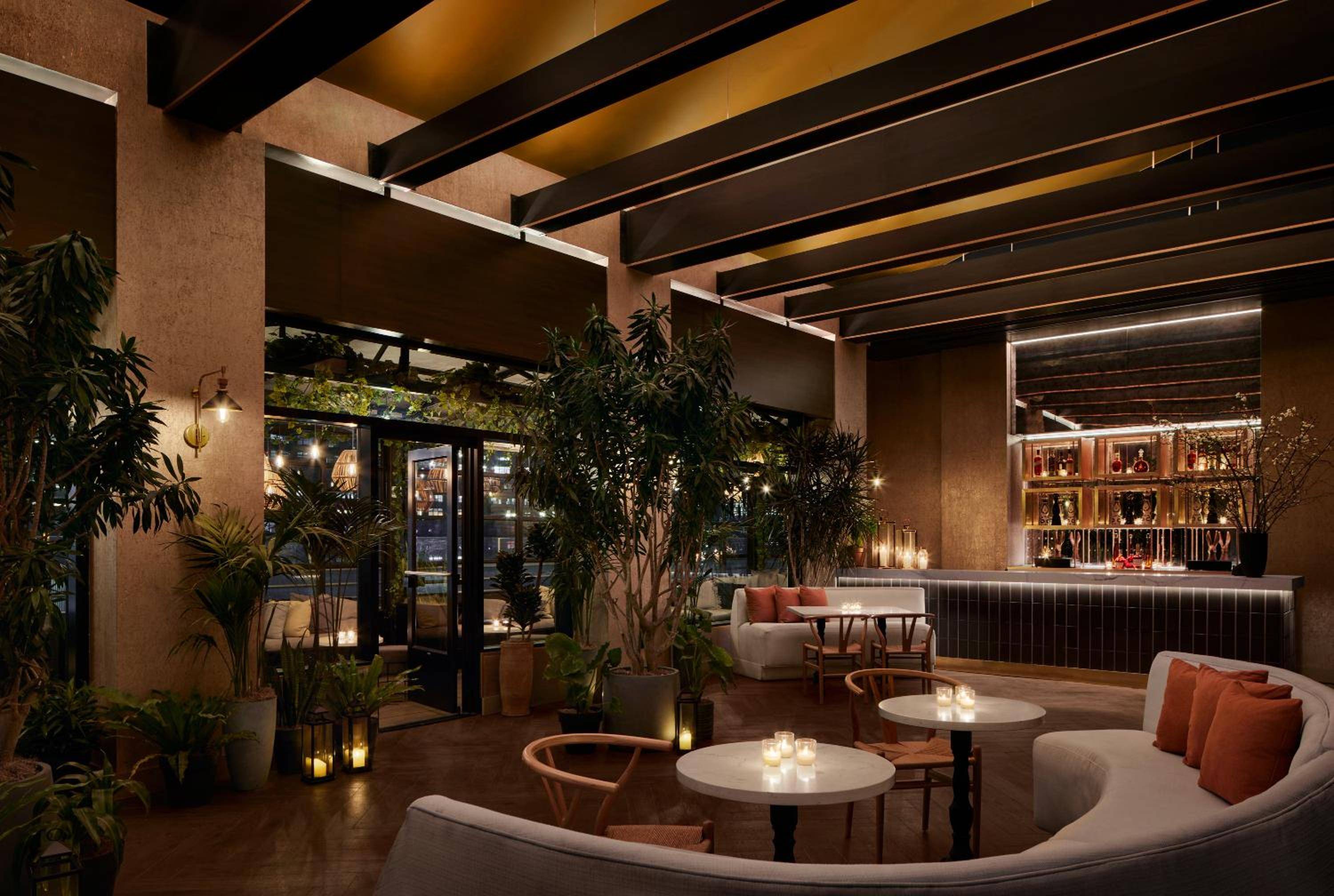 Candlelit tables beneath exposed wooden beams create intimate conversation nooks, while lush tropical plants soften the industrial-chic rooftop bar's sophisticated edge.