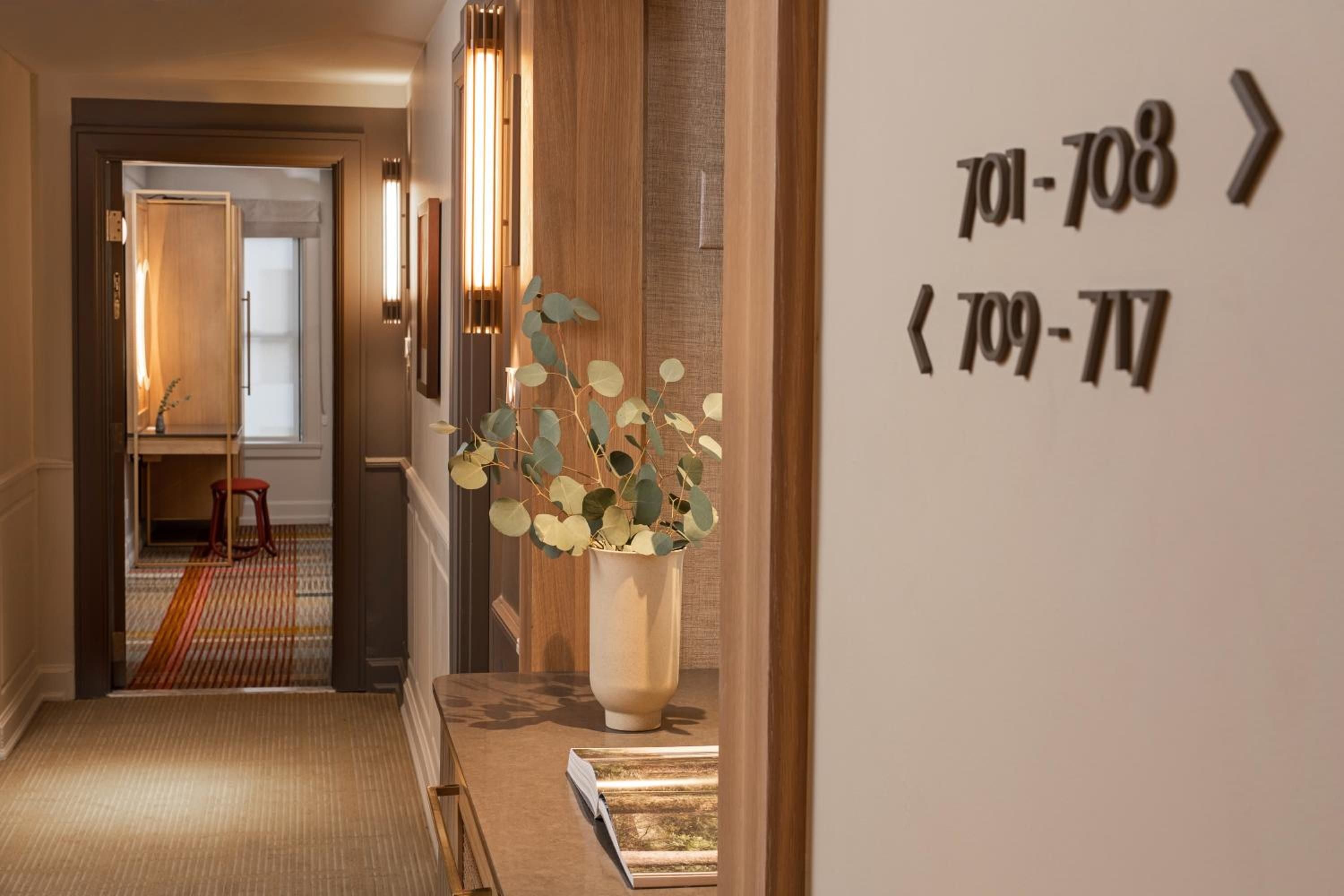 Warm wood paneling and brass sconces create an intimate corridor atmosphere, where eucalyptus arrangements and geometric carpeting guide guests toward thoughtfully numbered suites.