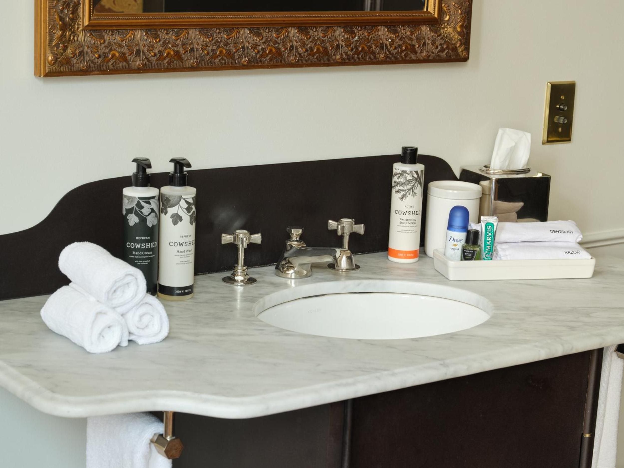Marble vanities gleam with brass fixtures and premium Cowshed amenities, while plush white towels and thoughtful touches create an atmosphere of refined luxury.