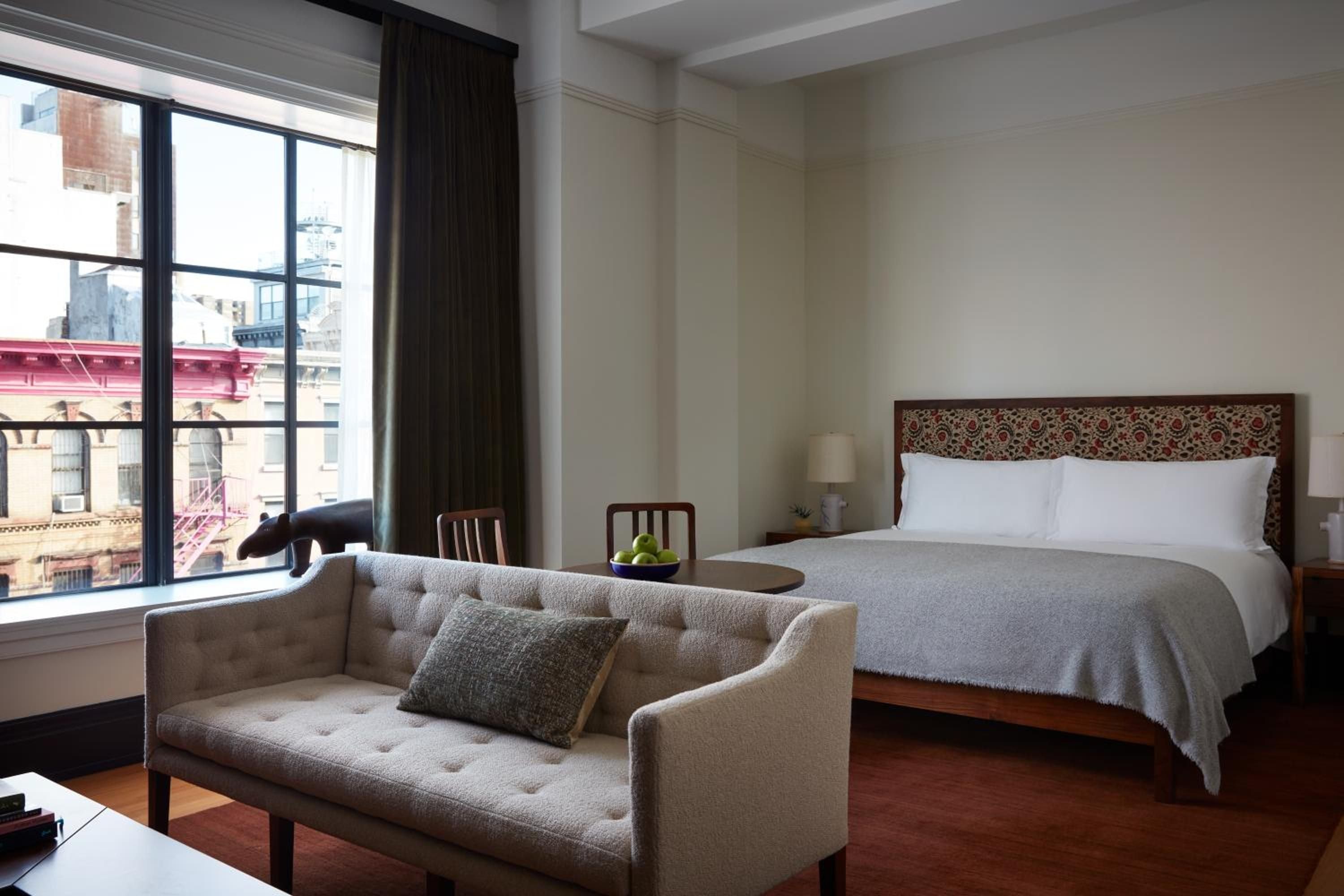 Floor-to-ceiling windows frame Lower East Side fire escapes while a tufted sofa and patterned headboard create intimate sanctuary above polished hardwood floors.