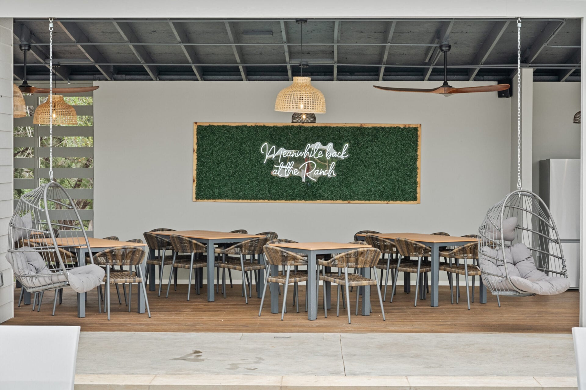 Woven rattan chairs and suspended egg seats create an inviting dining pavilion where rustic ranch charm meets contemporary comfort beneath warm pendant lighting.