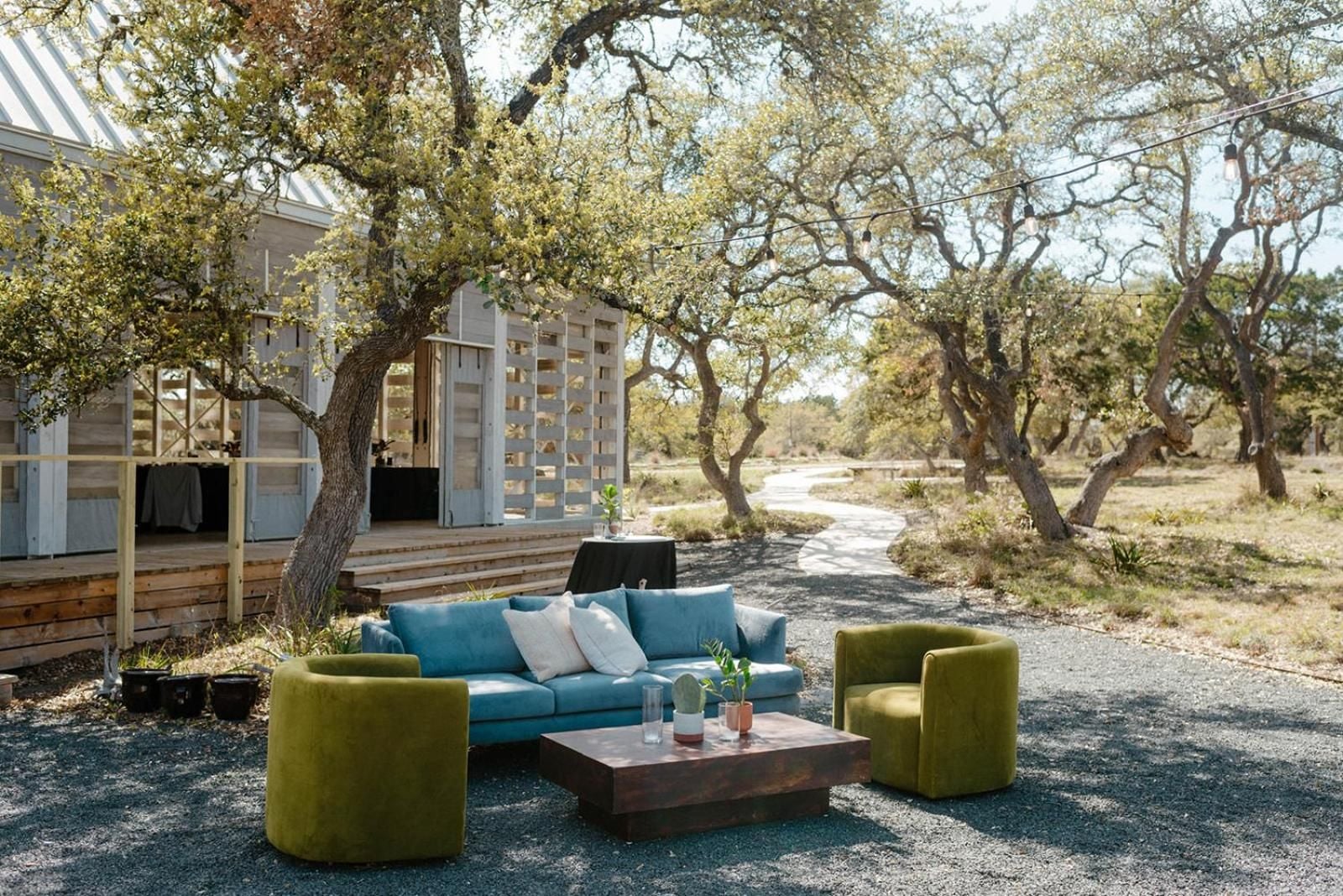Weathered oak trees shelter an unexpected outdoor lounge where teal cushions and chartreuse chairs create a desert oasis beneath dappled sunlight and rustic ranch architecture.