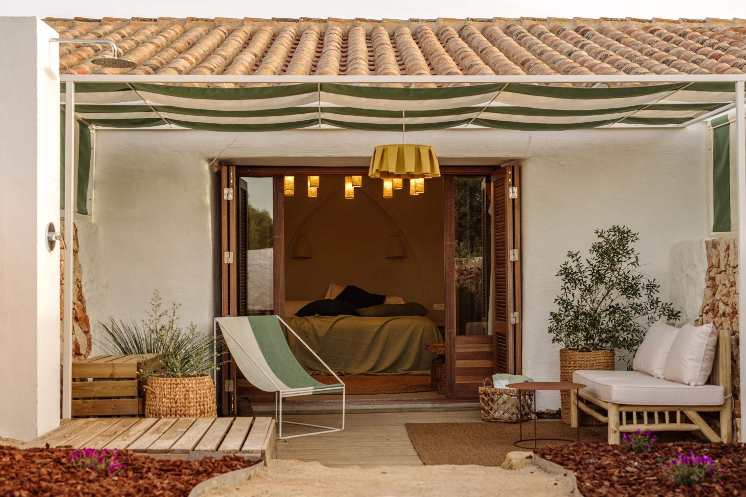 Folding wooden doors dissolve the boundary between bedroom and terrace, where Mediterranean tiles frame intimate conversations under striped canvas shade and golden pendant light.