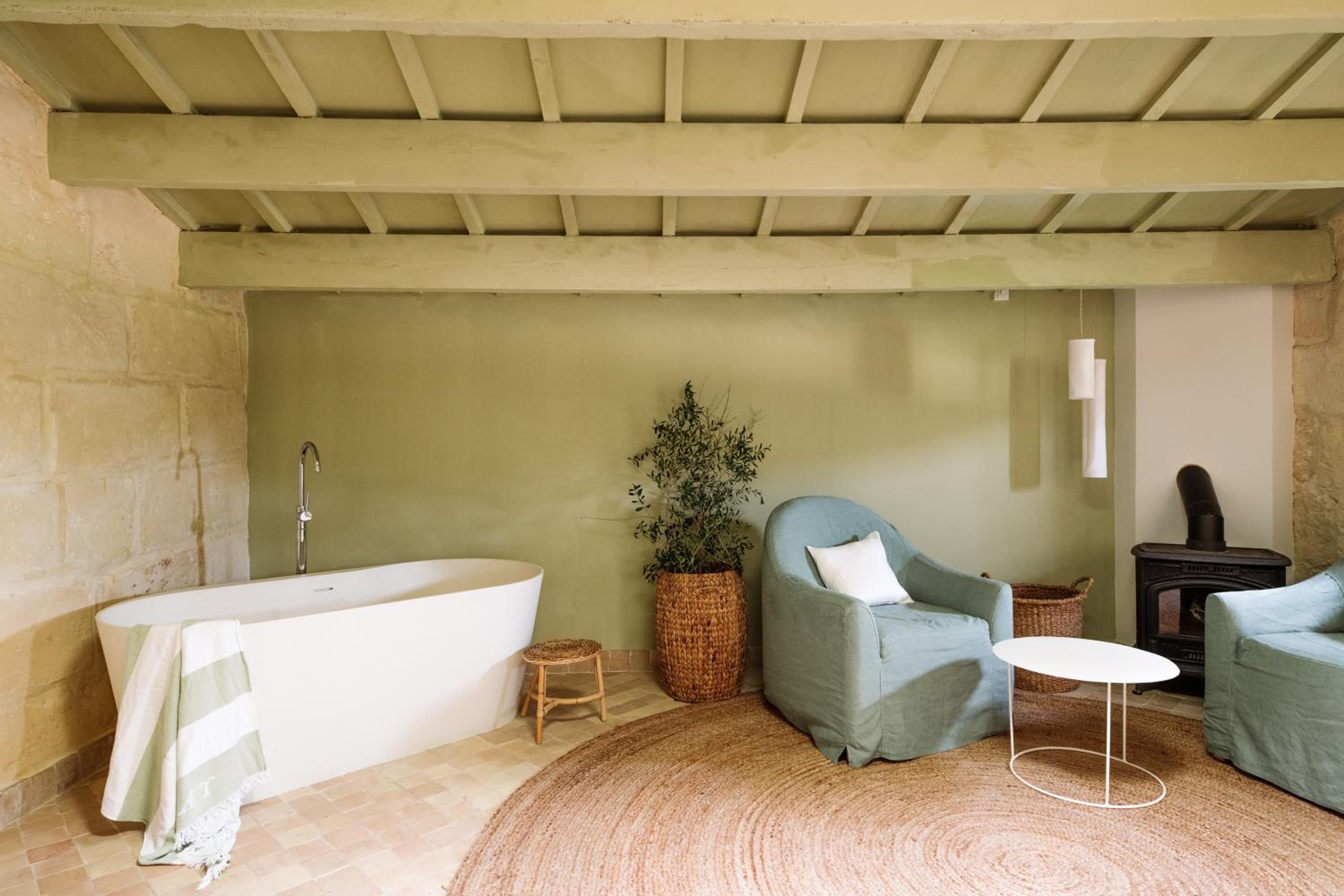 Exposed stone beams frame this serene bathroom suite where sage-green armchairs invite lingering beside the sculptural freestanding tub and crackling wood stove.
