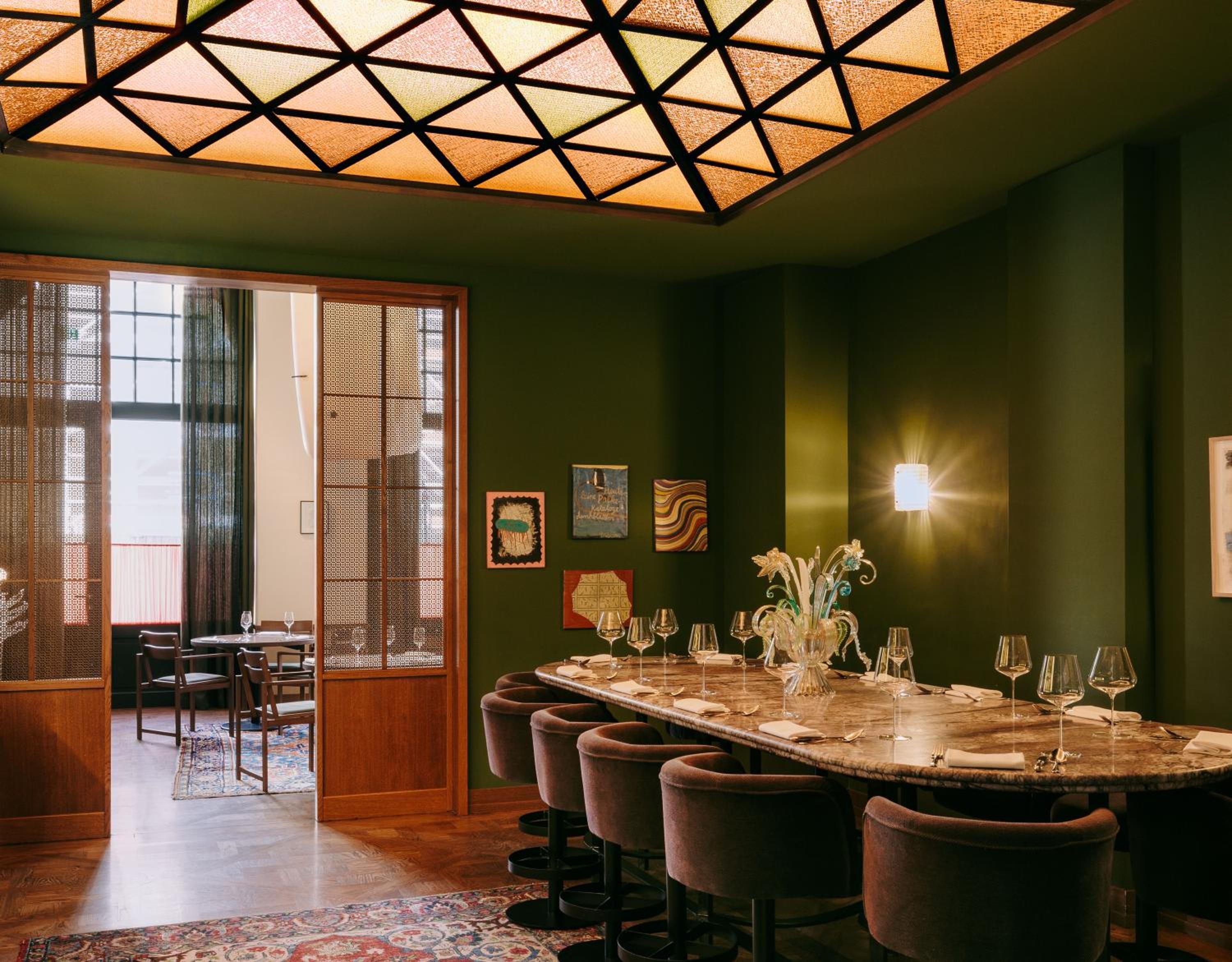 Rich olive walls frame an intimate dining sanctuary where geometric skylights cast honeyed light across marble tables and sumptuous velvet chairs, creating sophisticated warmth.