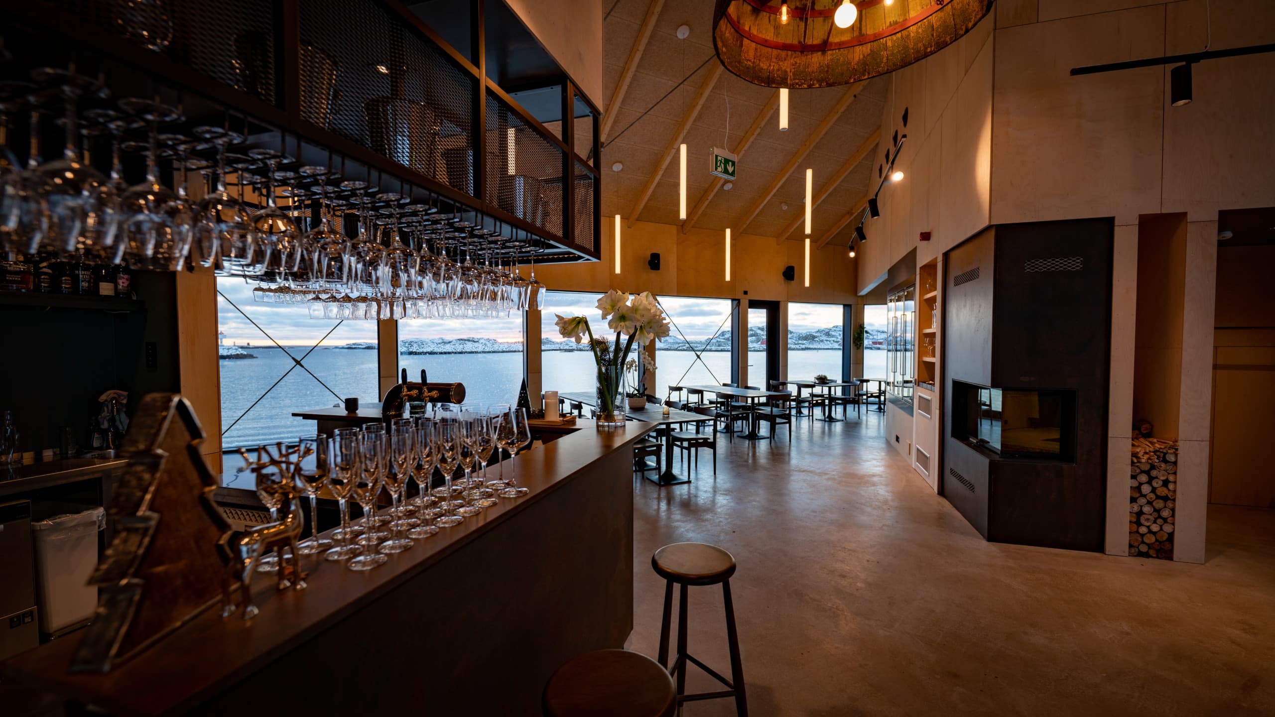 Floor-to-ceiling windows frame Arctic waters while suspended wine glasses catch golden light above the sleek bar, creating an intimate Nordic dining sanctuary.
