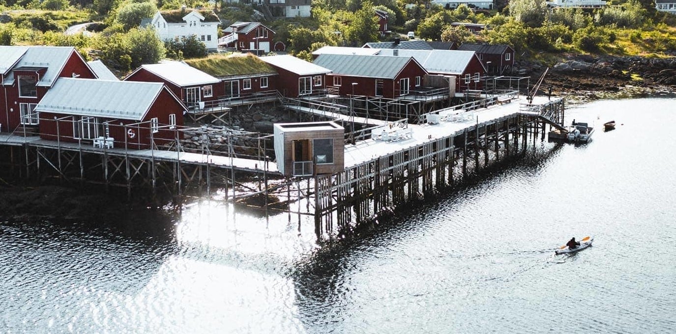 Traditional red fishermen's cabins perch on weathered stilts above crystalline waters, where kayakers glide beneath the wooden pier connecting this authentic Norwegian lodge to the sea.
