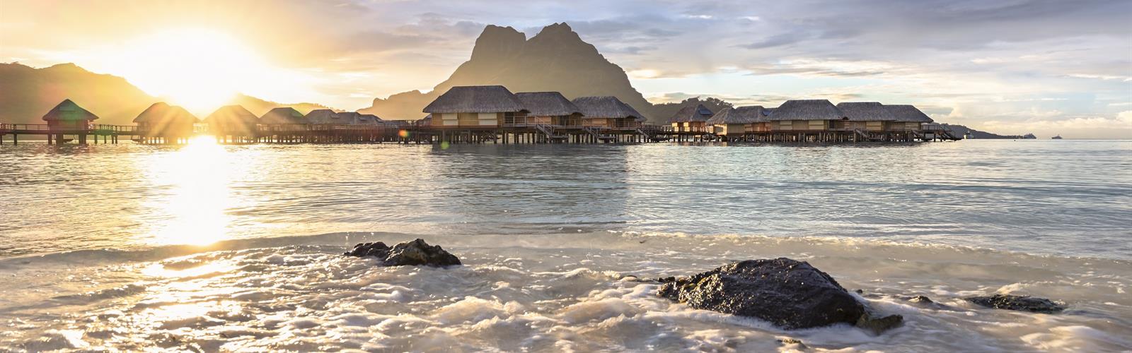 Golden hour bathes Bora Bora's iconic overwater bungalows in ethereal light, while gentle waves lap against volcanic rocks on this pristine Polynesian shore.
