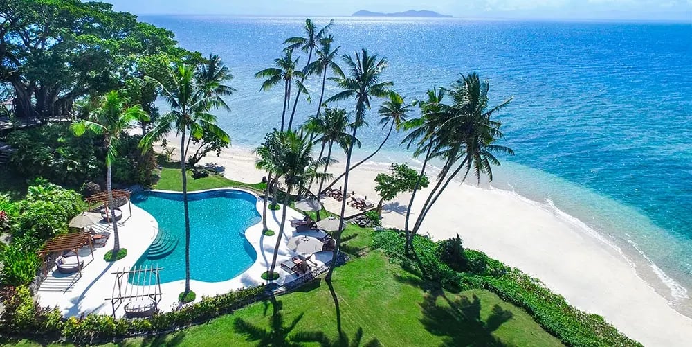 Turquoise infinity pool curves along pristine white sand where swaying coconut palms frame azure waters, creating an intimate tropical sanctuary on this private Fijian isle.