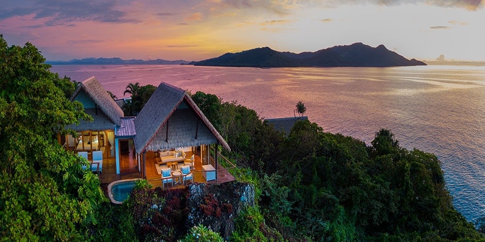 Thatched-roof villas perch dramatically above turquoise waters, their private pools catching the golden light as volcanic peaks dissolve into Fiji's endless Pacific horizon.