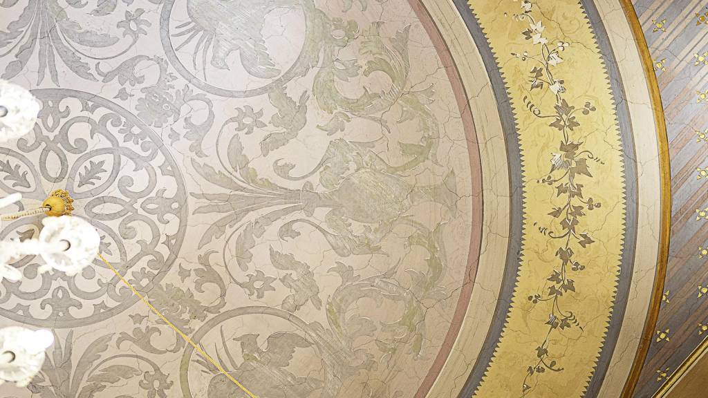 Ornate ceiling frescoes reveal centuries of Italian craftsmanship, where gilded botanical motifs dance across vaulted surfaces in muted gold and sage green palettes.