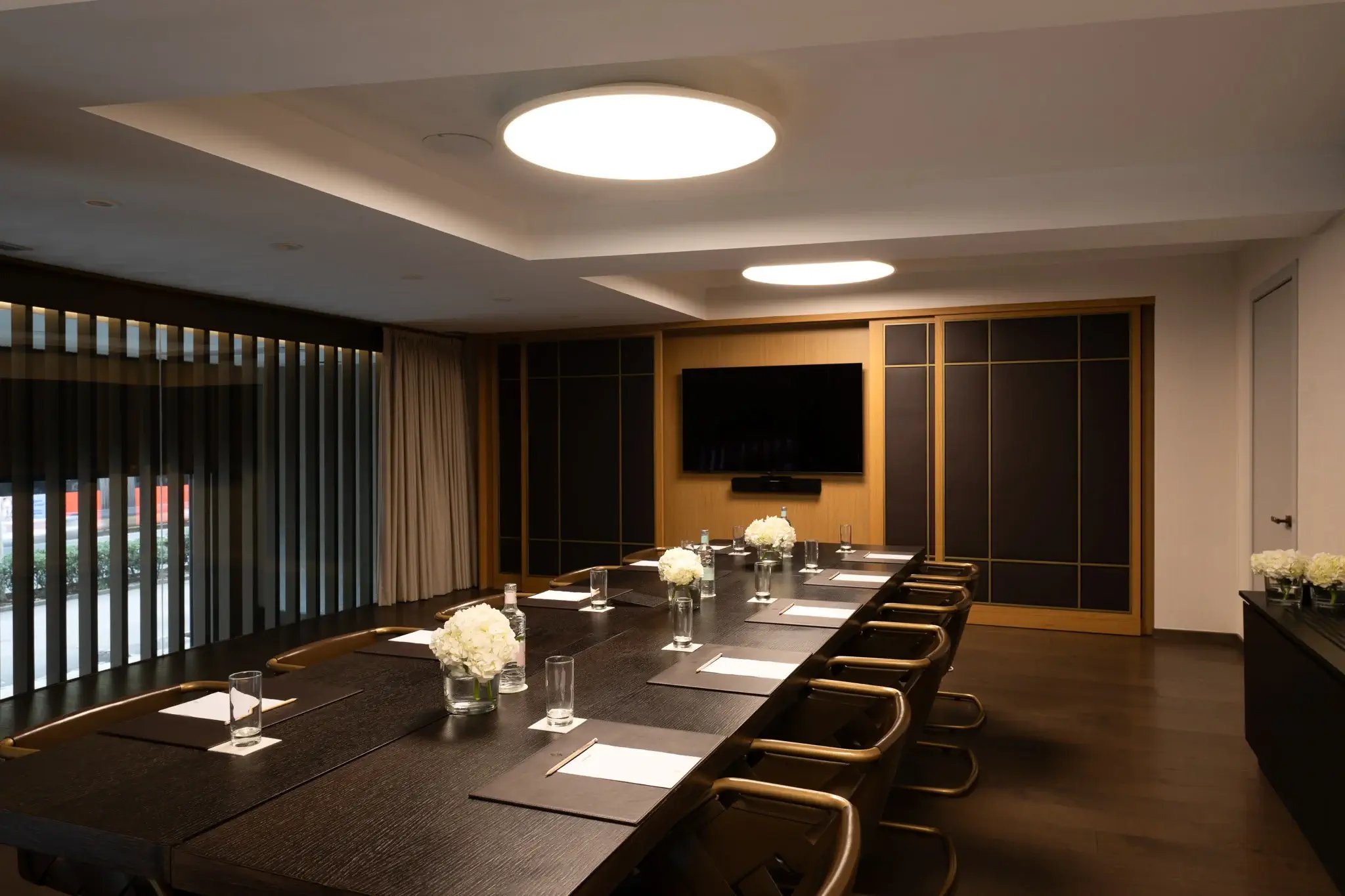 Warm wood paneling and sleek black furnishings create an intimate boardroom where natural light filters through vertical blinds, elevating business gatherings beyond the ordinary.