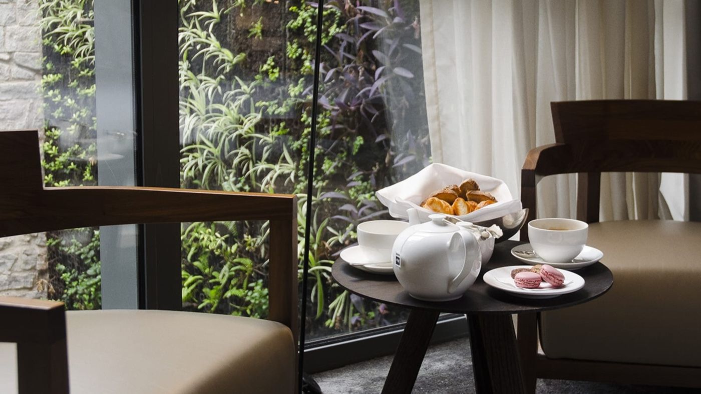 Morning light filters through floor-to-ceiling windows as a perfectly arranged tea service awaits on sleek dark wood, overlooking the hotel's verdant living wall.