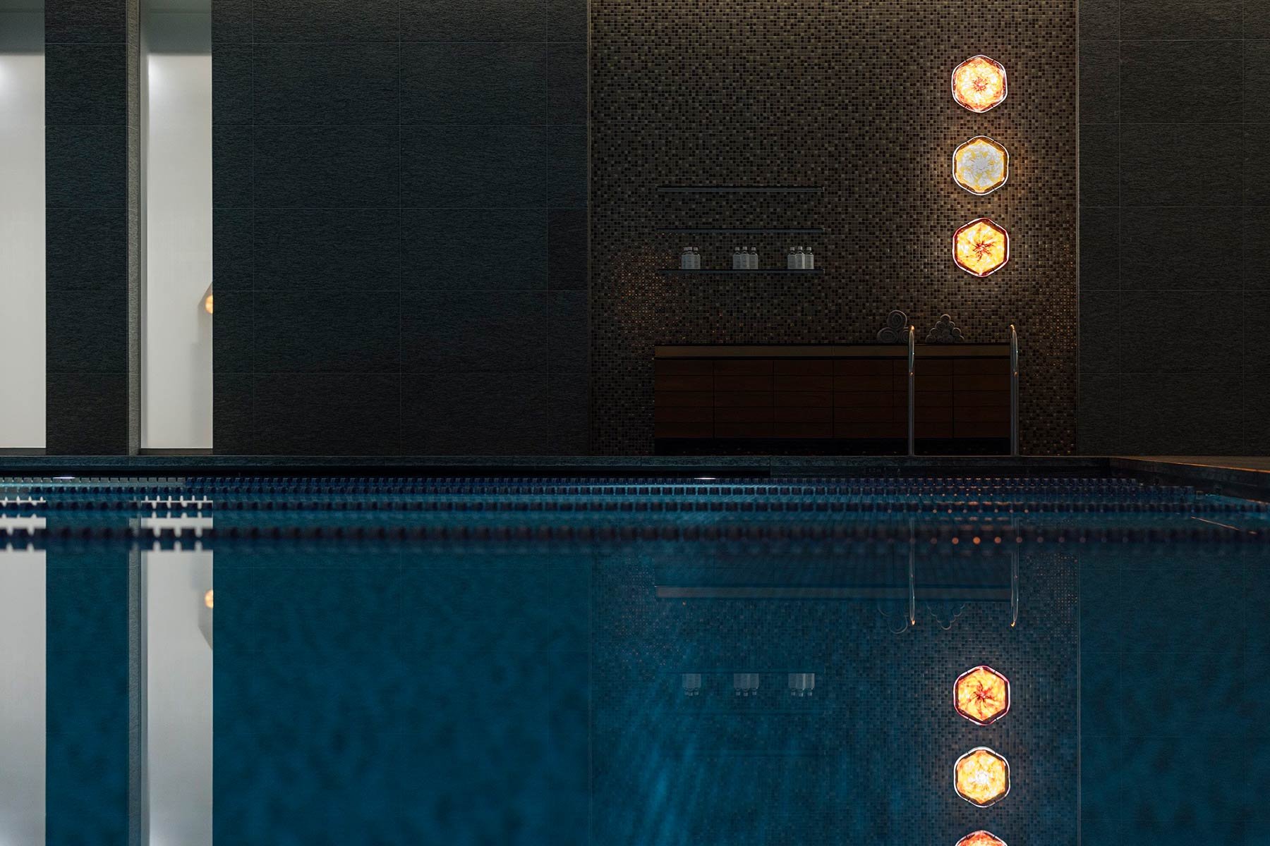 Hexagonal art pieces glow like lanterns against charcoal mosaic walls, their warm light dancing across the indoor pool's mirror-still surface in perfect symmetry.