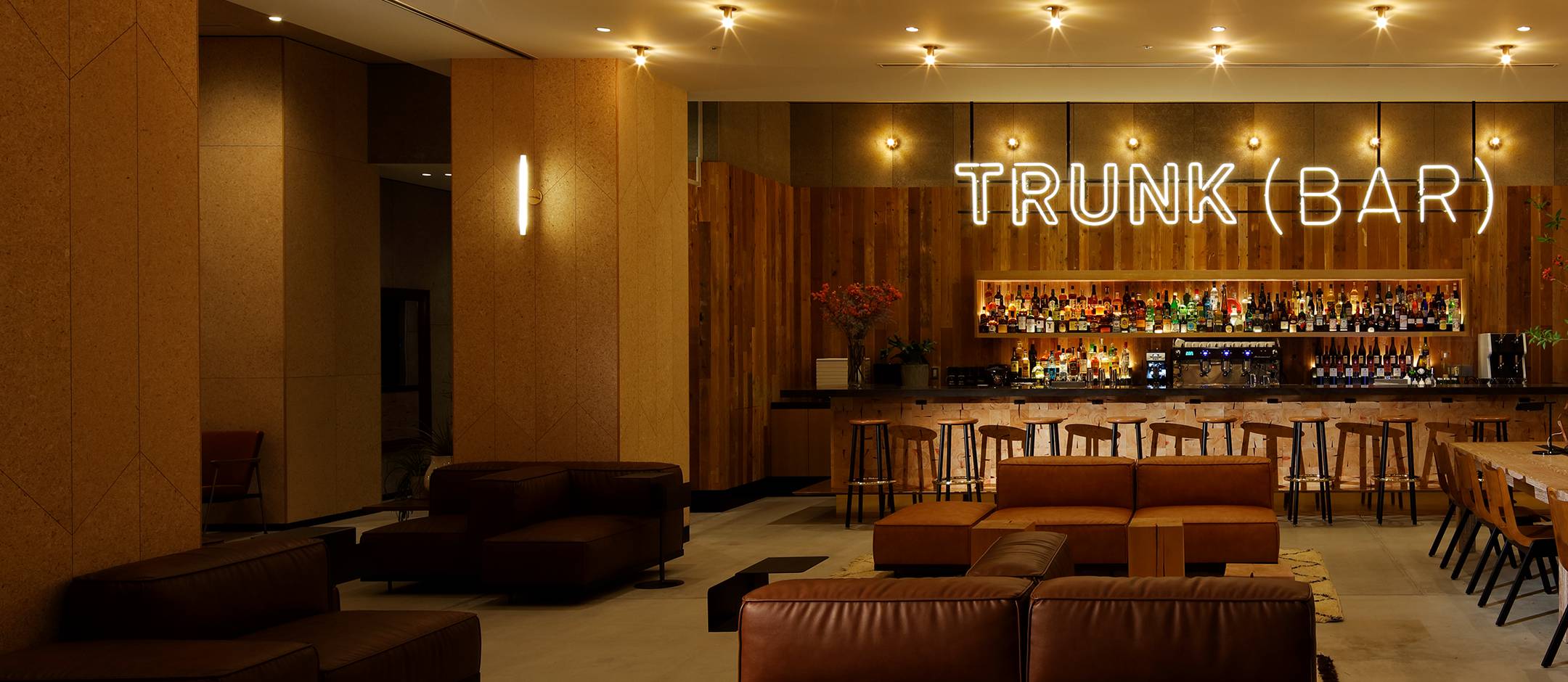 Warm wood panels and rich leather seating create an intimate atmosphere at TRUNK(BAR), where neon signage glows against curated spirits and soft ambient lighting.