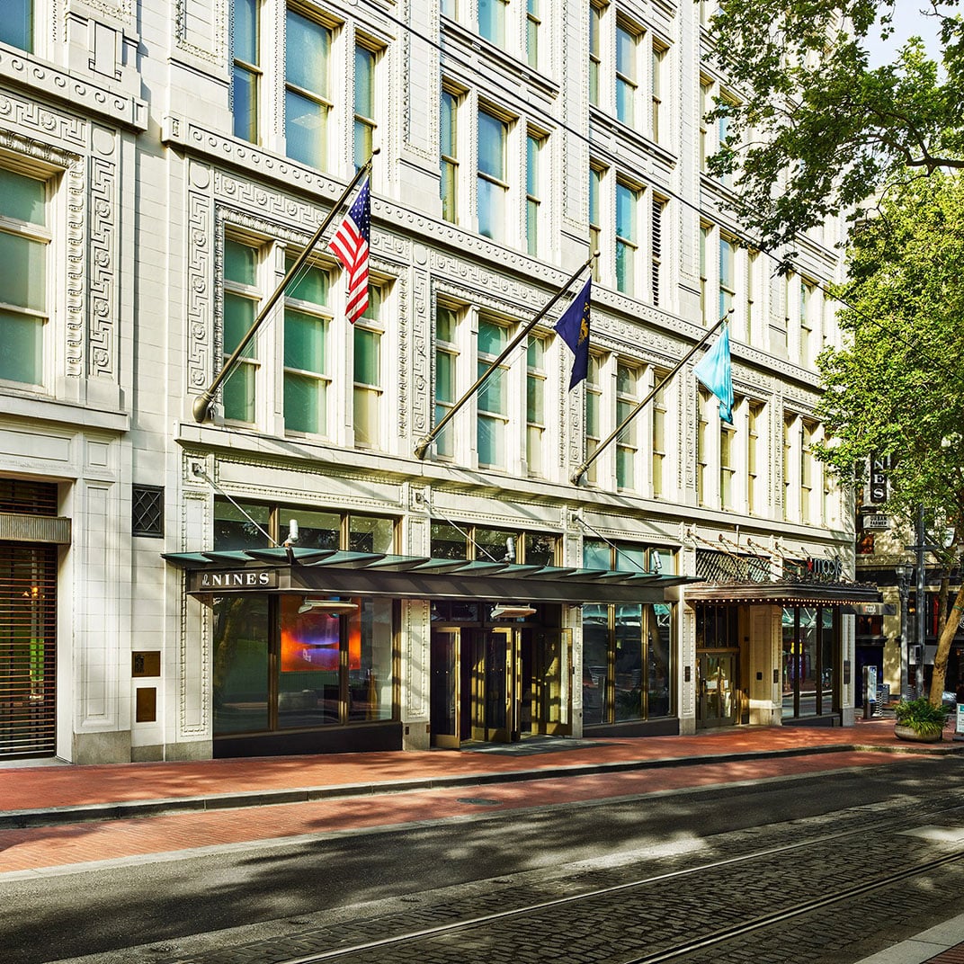 Historic Meier & Frank building's ornate terra cotta facade gleams in afternoon light, where brass revolving doors welcome guests beneath ceremonial flags on downtown Portland's bustling streets.