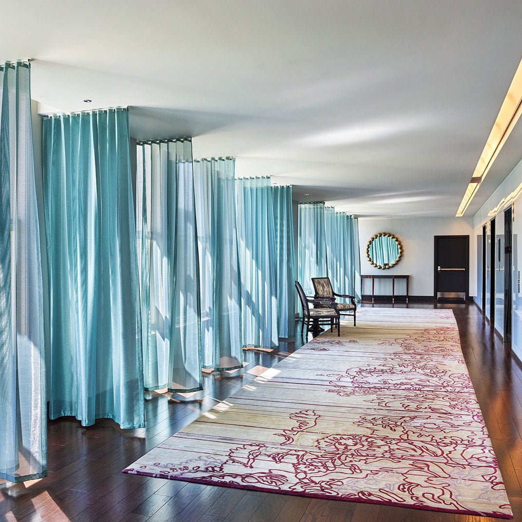 Flowing turquoise curtains create an undulating corridor of color, where natural light dances across polished floors and intricate carpet patterns guide guests through this sophisticated passage.