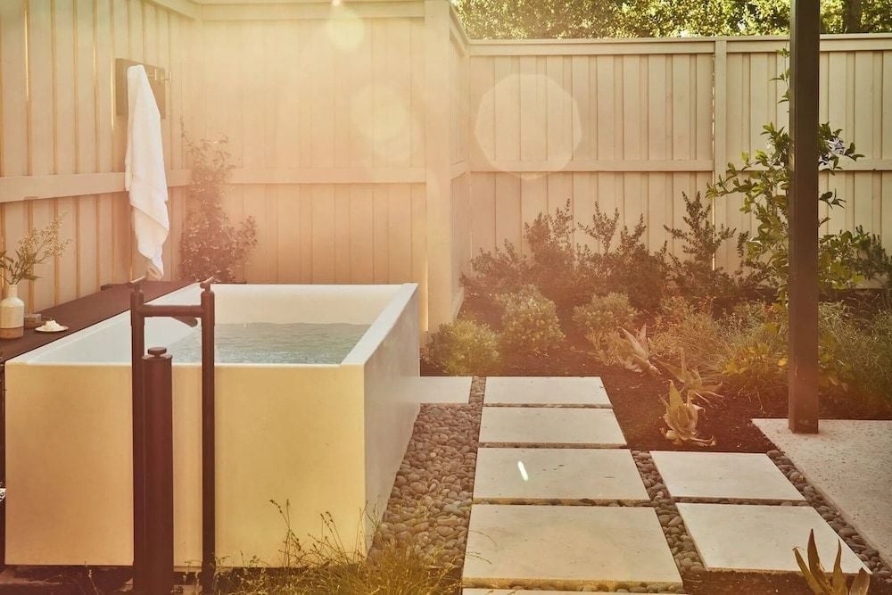 Golden hour light filters through steam rising from a private soaking tub, where smooth stone pathways wind through native plantings in this serene outdoor spa retreat.