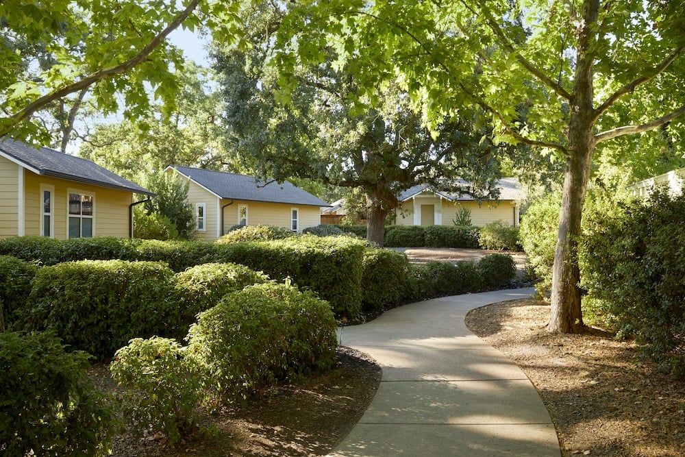 Meandering pathways wind through meticulously manicured gardens, connecting intimate cottage-style accommodations beneath towering shade trees that dapple the walkways with golden afternoon light.
