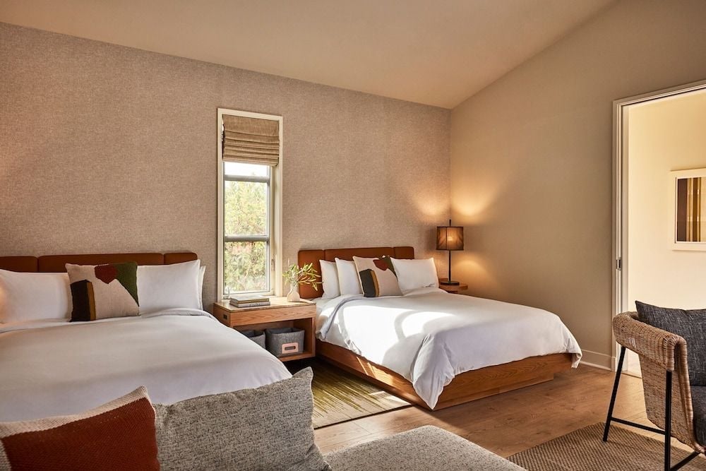 Twin beds dressed in crisp white linens anchor this serene guest room, where natural textures and warm lighting create an intimate sanctuary overlooking Calistoga's rolling landscape.