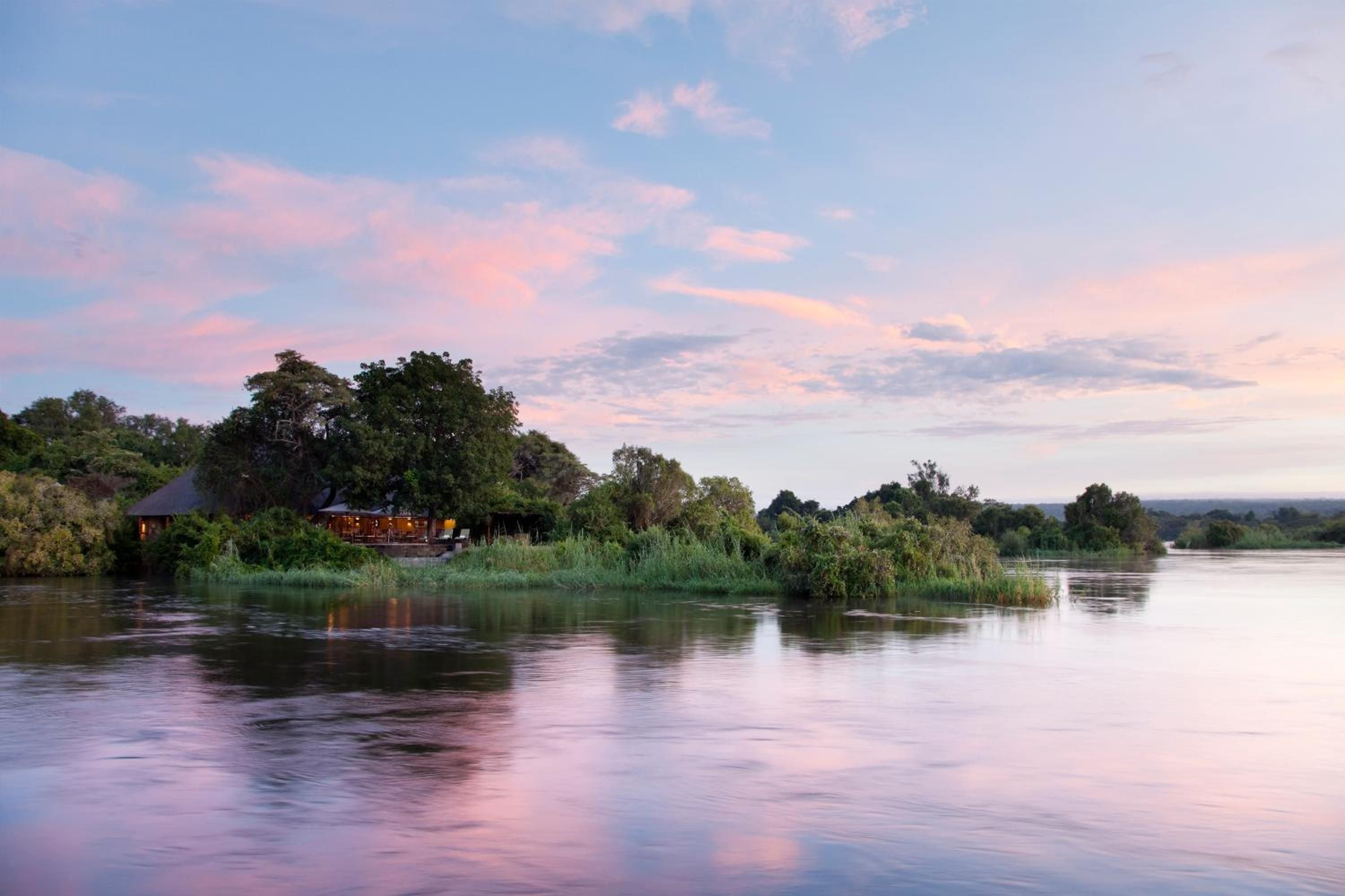 Sunset paints the Zambezi in rose gold as Royal Chundu's thatched pavilion glows warmly among ancient trees, creating an intimate African riverside sanctuary.