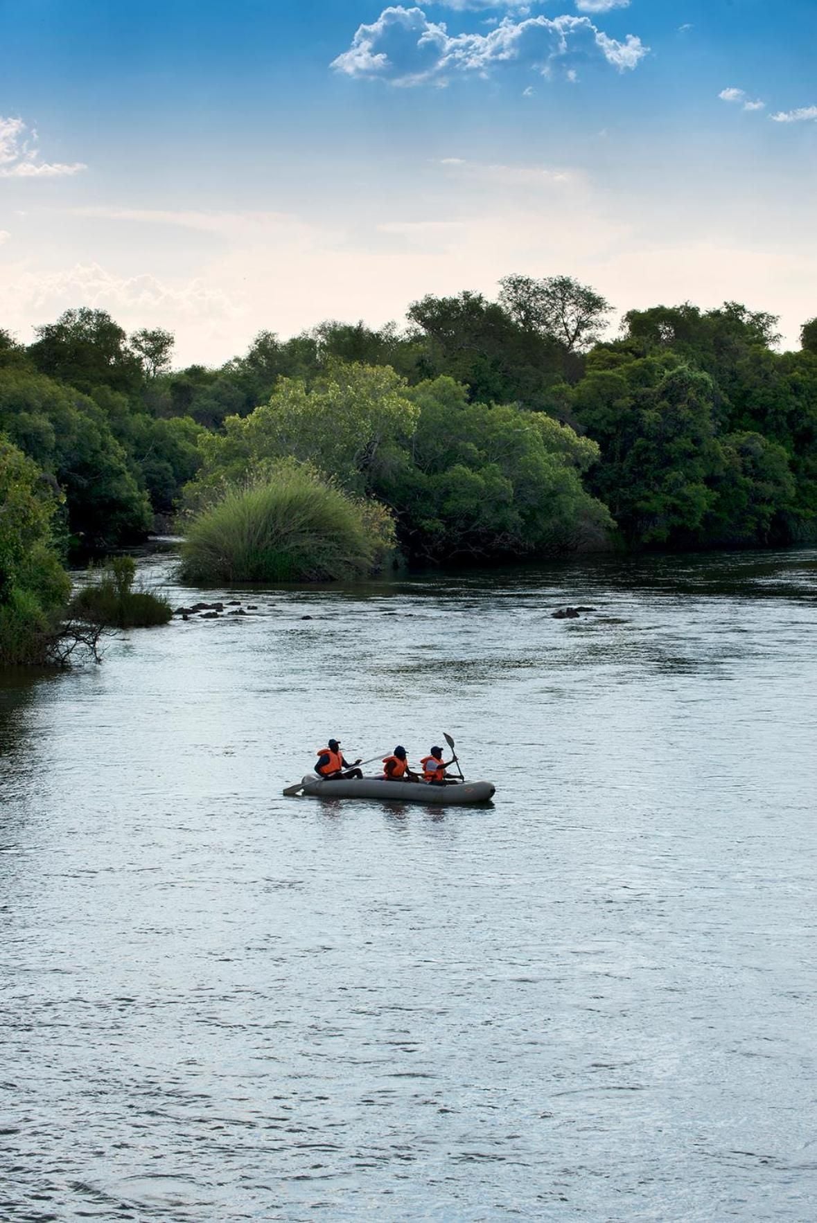 Gentle rapids ripple beneath towering riverine forest as guests drift downstream in inflatable rafts, paddles cutting through the Zambezi's sun-dappled current.