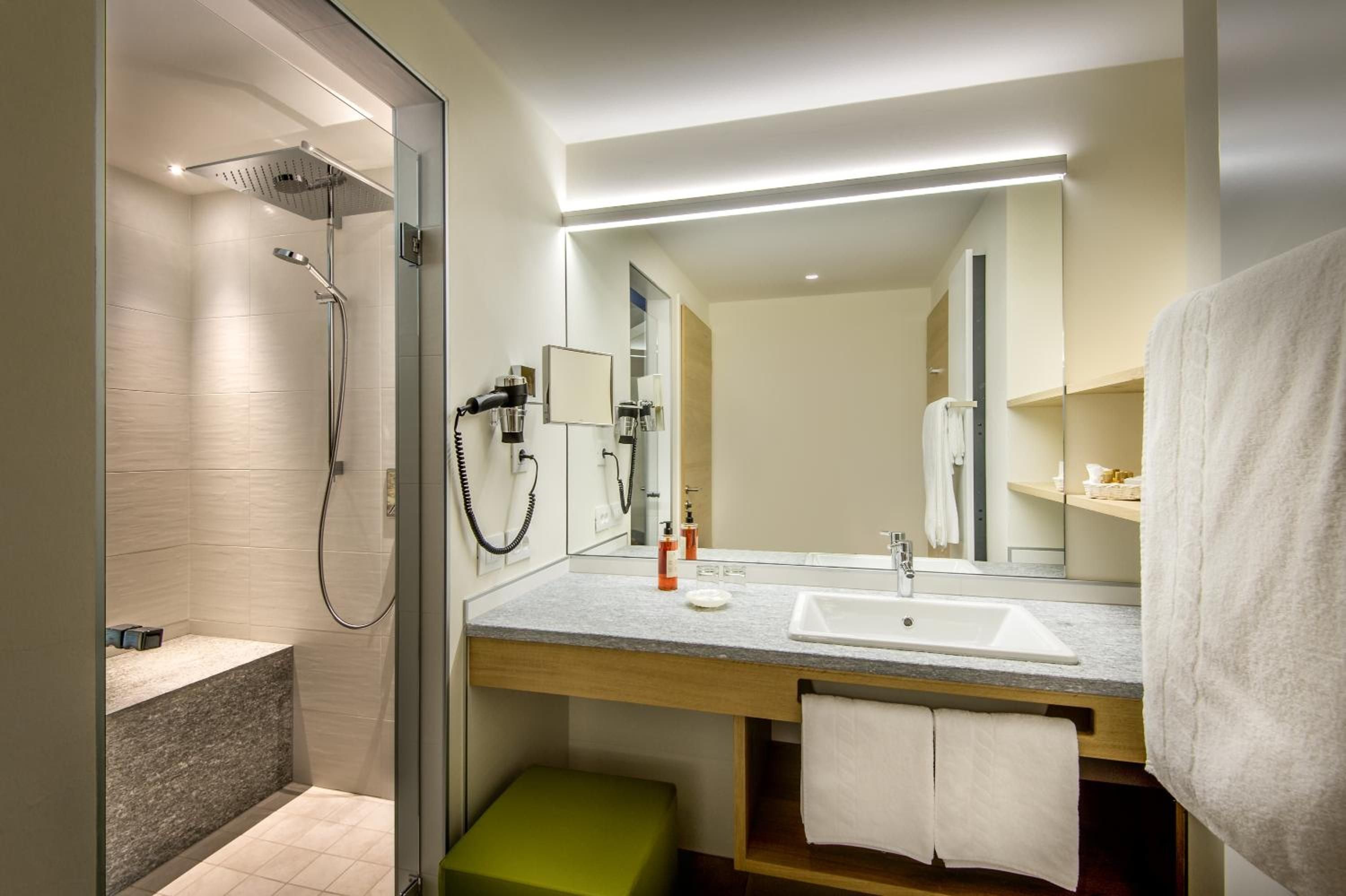 Rain shower and illuminated vanity create a serene sanctuary, where warm wood tones meet cool marble beneath soft LED lighting that whispers luxury.