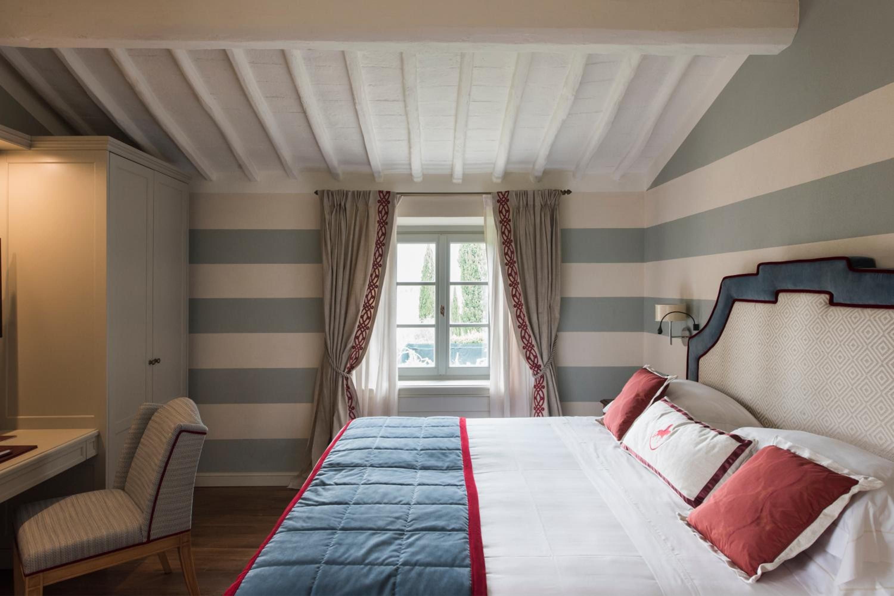 Exposed white beams crown this elegant guest room where sage stripes meet coral accents, creating rustic Tuscan charm with sophisticated flair.