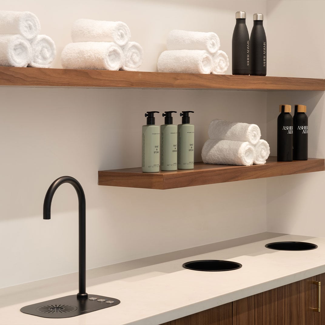 Warm wood shelving displays pristine rolled towels and premium amenities above sleek treatment stations, where matte black fixtures create an unexpectedly sophisticated spa sanctuary.