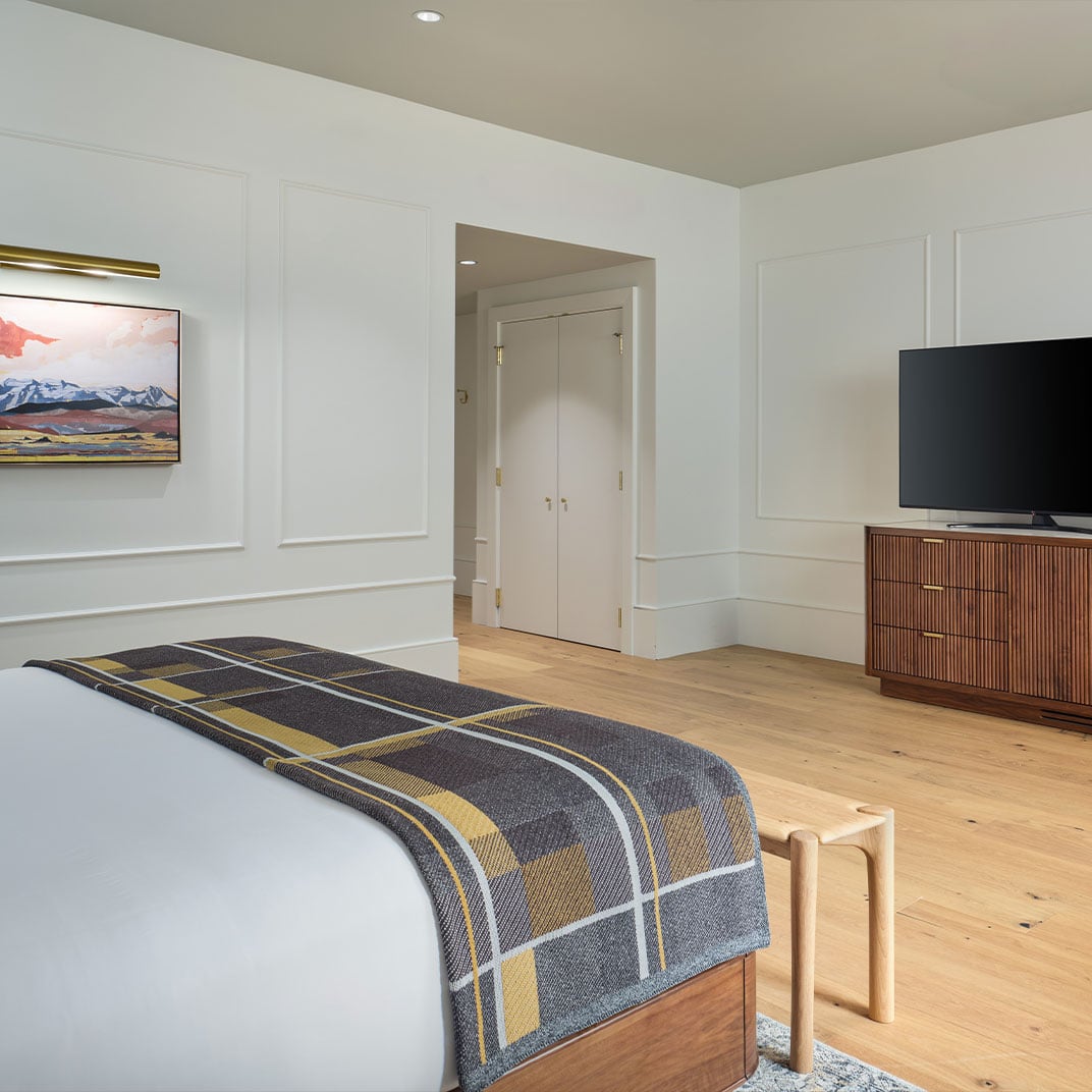 Warm wood floors meet crisp white wainscoting in this thoughtfully appointed guest room, where plaid wool throws and brass accents create mountain lodge sophistication.