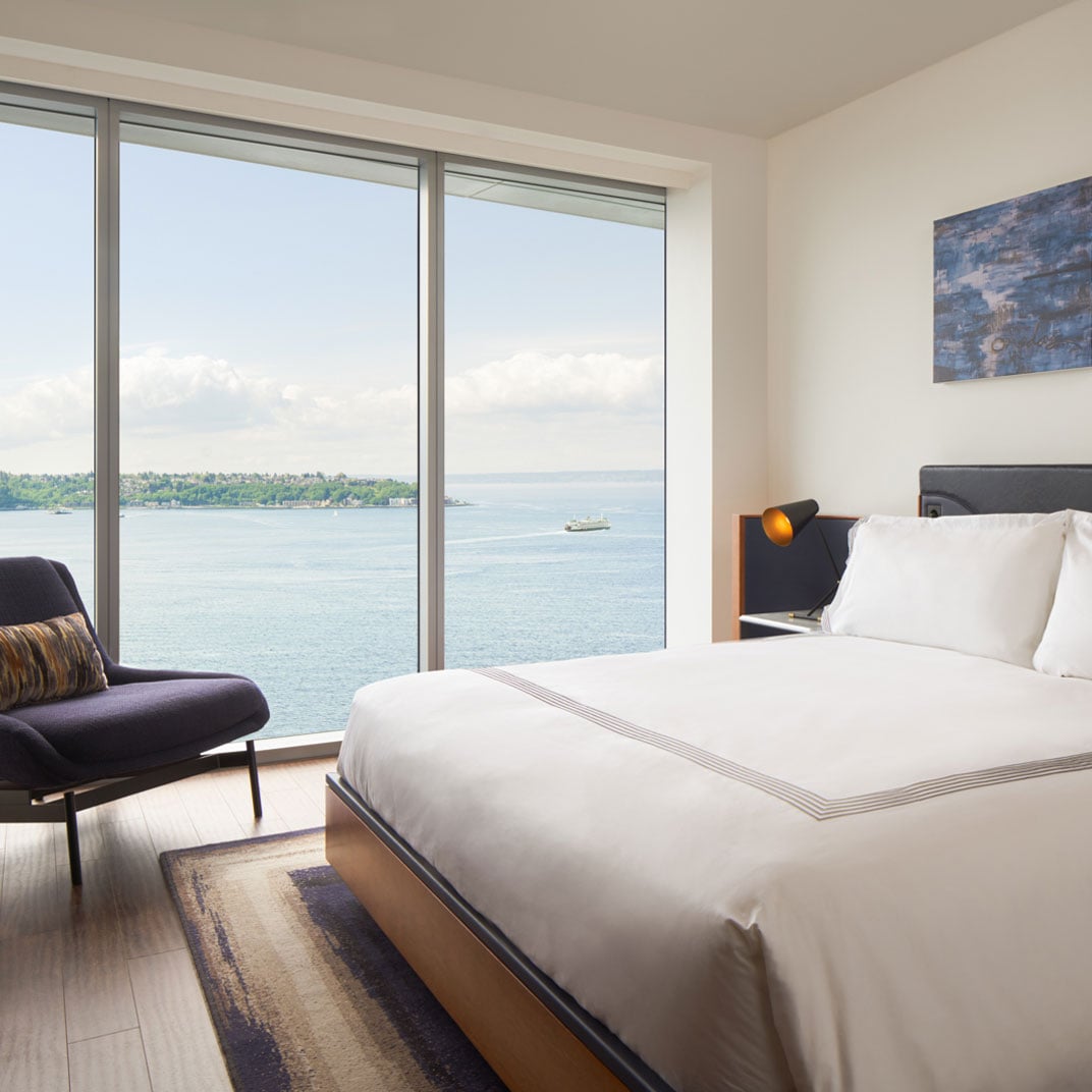 Floor-to-ceiling windows frame Elliott Bay's shimmering waters from this waterfront suite, where crisp white linens meet warm wood tones and purple accent chairs.