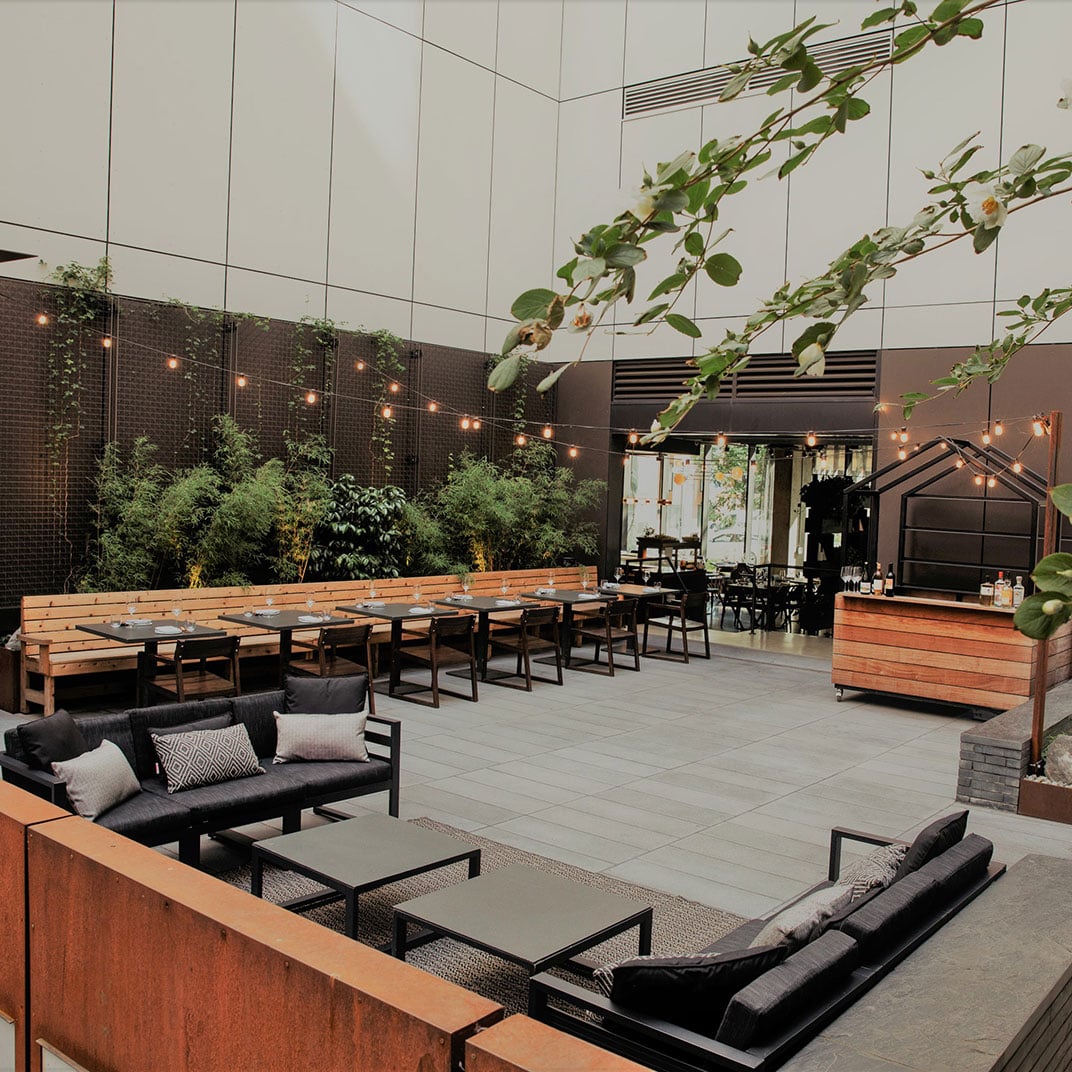 String lights illuminate the rooftop terrace where modern lounge seating mingles with dining tables beneath overhead greenery and dramatic urban architecture.