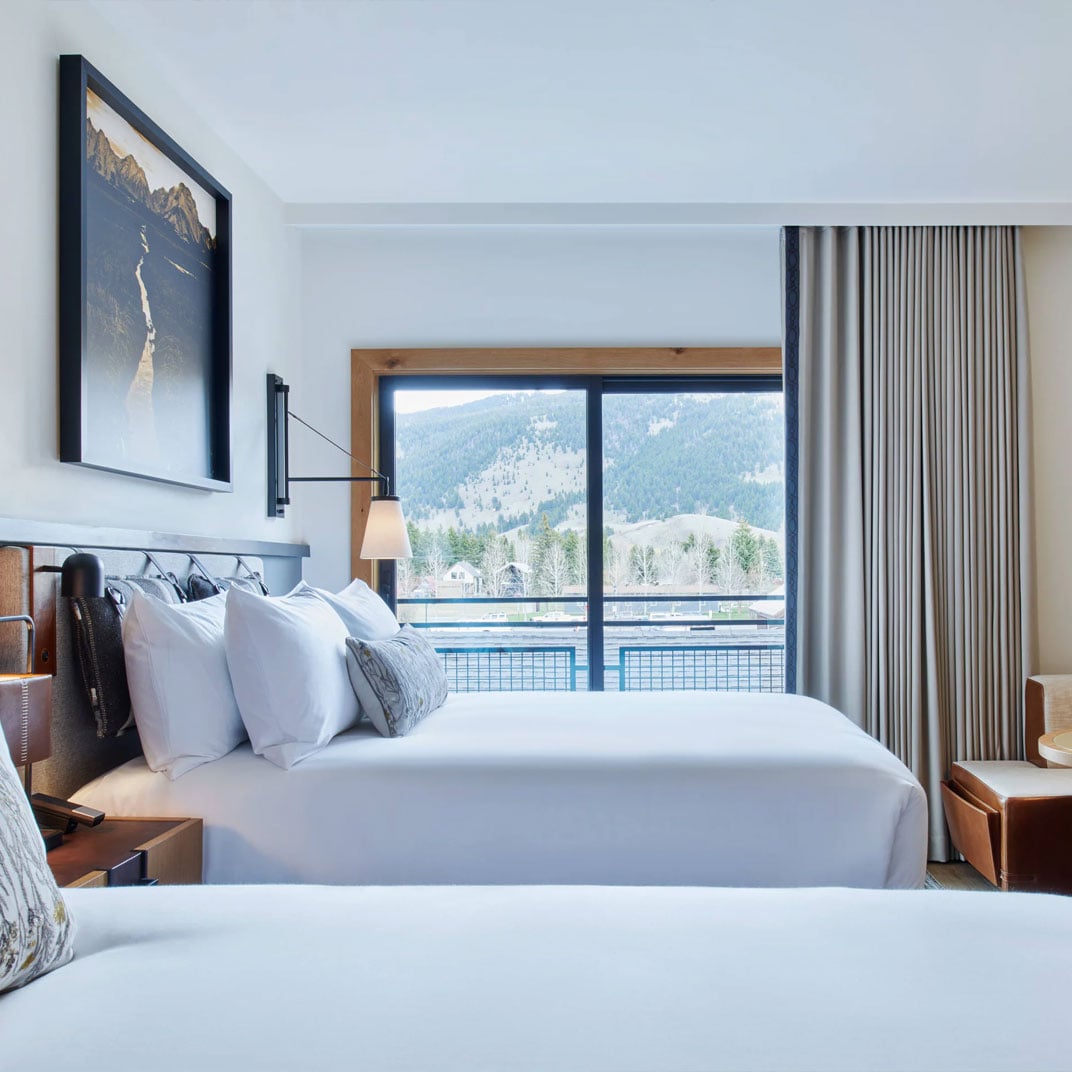 Crisp white linens frame floor-to-ceiling windows revealing Jackson Hole's dramatic mountain backdrop, where modern alpine luxury meets untamed wilderness views.