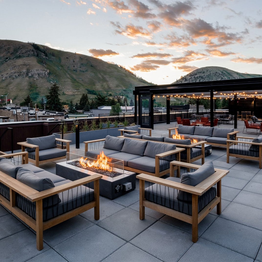 Fire pits crackle beneath string lights as the Teton Range glows pink at dusk from this sophisticated rooftop terrace with sleek teak seating.