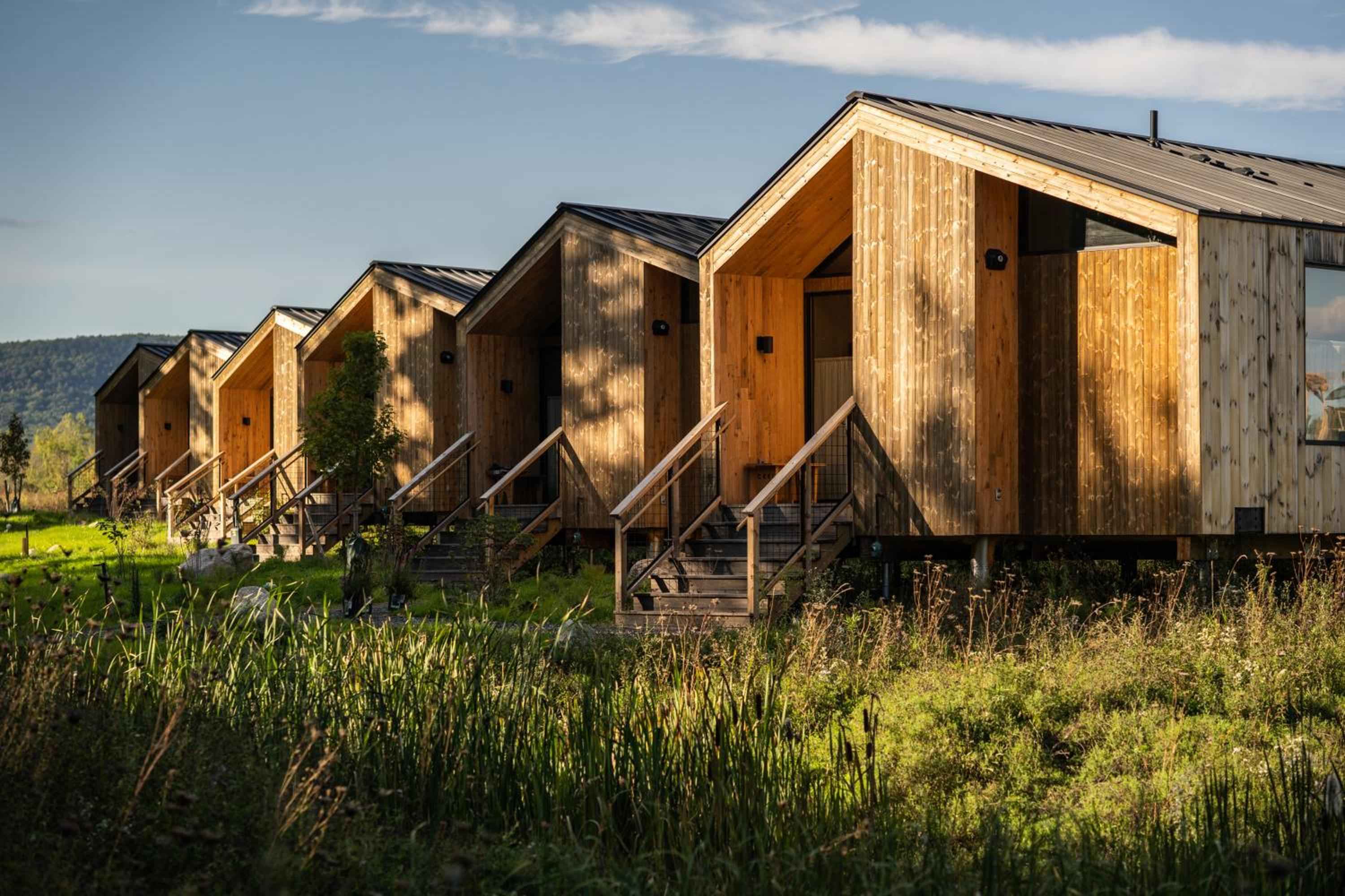 Elevated wooden cabins with weathered cedar siding create an intimate village feel, their warm honey tones glowing against rolling meadows dotted with wildflowers and native grasses.