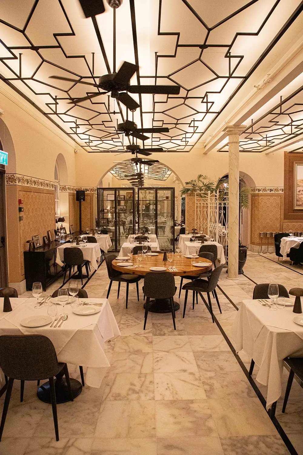 Geometric ceiling patterns cast dramatic shadows over intimate dining tables, where modern black chairs meet crisp white linens beneath ornate archways and warm marble floors.