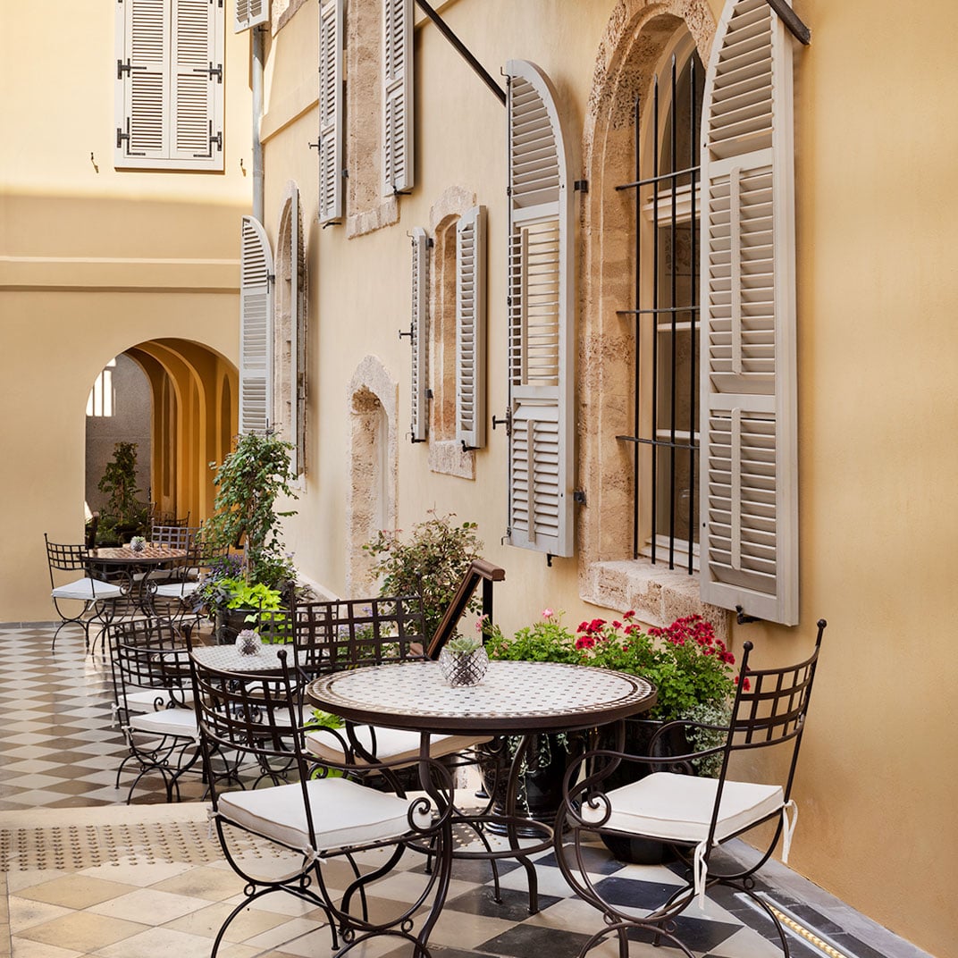 Wrought-iron café tables dot the sun-drenched courtyard, where weathered limestone walls and white shutters create an intimate Mediterranean refuge steps from bustling Tel Aviv.