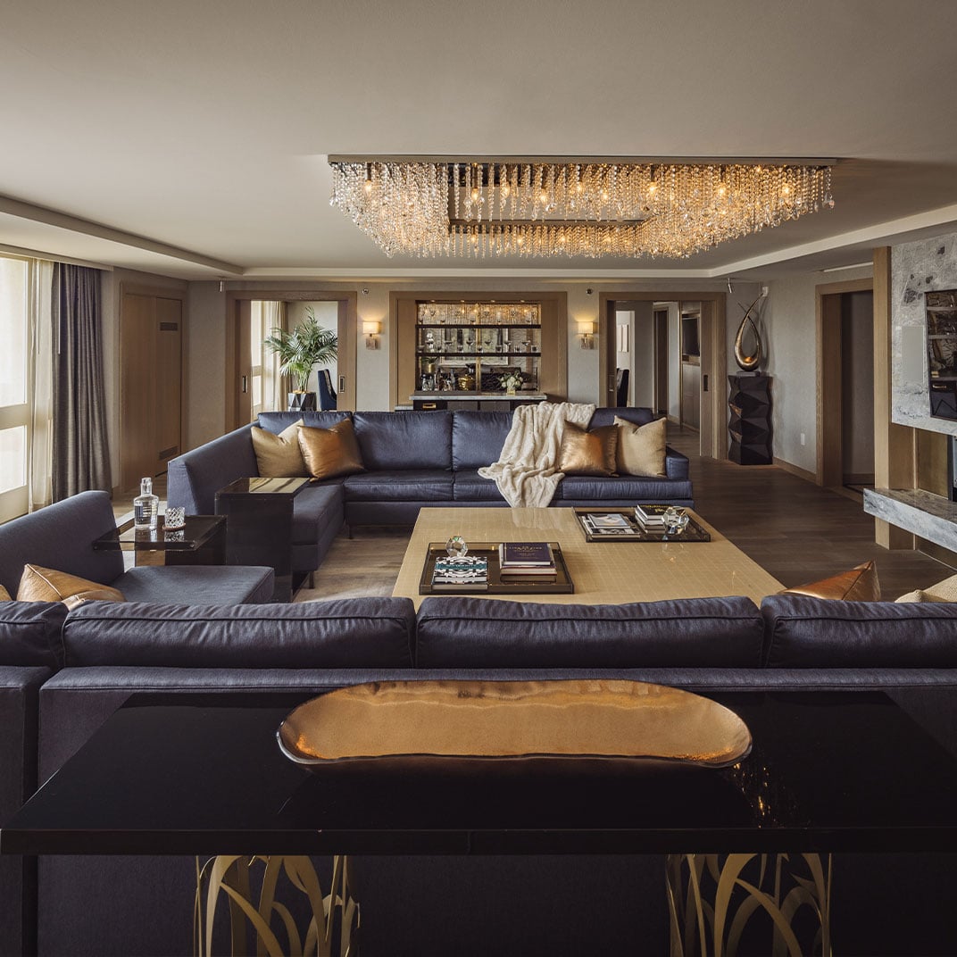 Crystal chandeliers cast warm light across navy leather sectionals and burnished gold accents in this penthouse suite, where contemporary luxury meets sophisticated comfort.