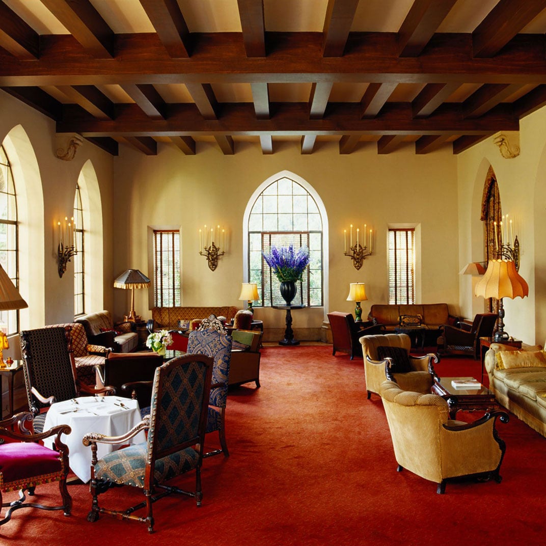 Exposed wooden beams frame this grand salon where jewel-toned Persian rugs meet gothic arches, creating an Old Hollywood refuge bathed in golden afternoon light.