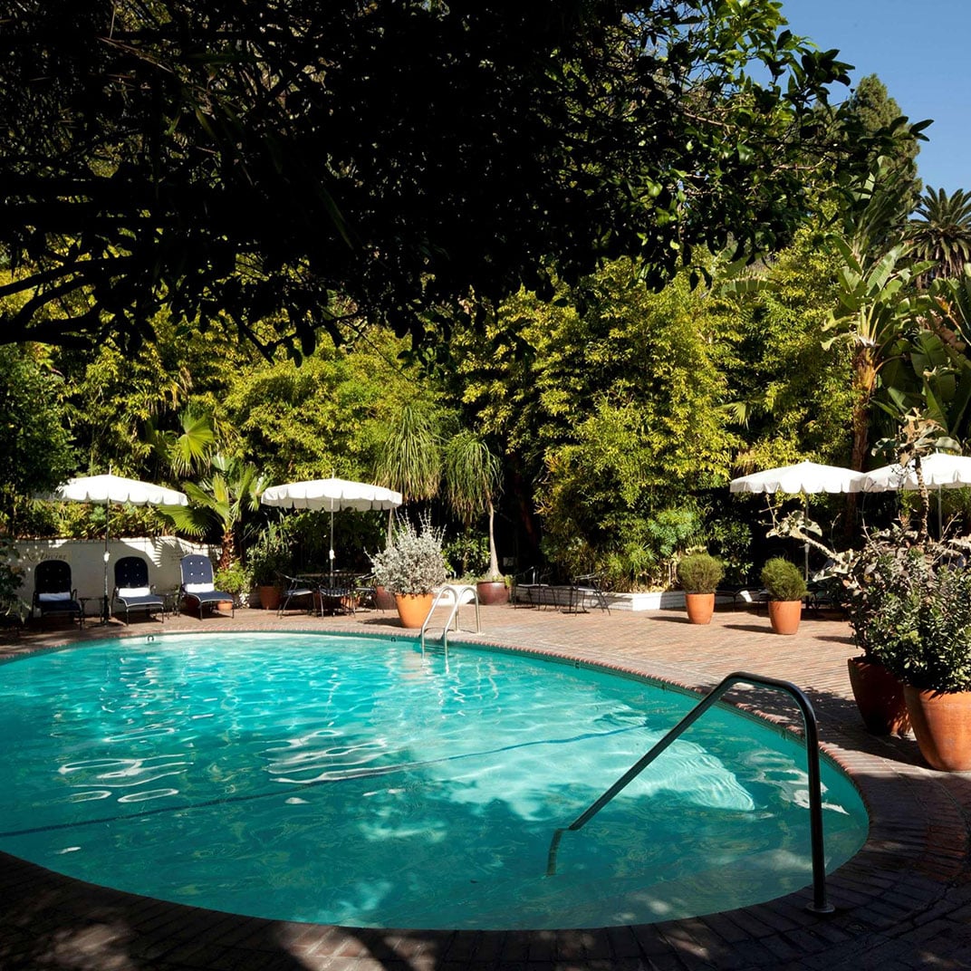 Dappled sunlight filters through towering palms and mature trees, casting dancing shadows across the secluded pool where white umbrellas offer refuge from Hollywood's golden afternoon light.