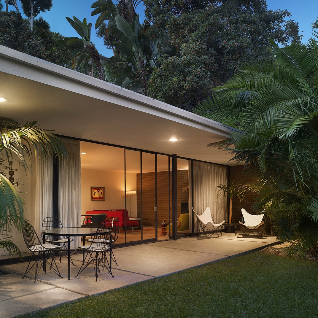 Mid-century modern lines dissolve into lush tropical foliage, where wire mesh chairs and butterfly seats create an intimate outdoor living room beneath the canopy.