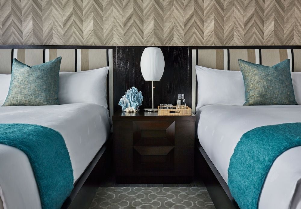 Twin beds dressed in crisp white linens and teal accents create a serene sanctuary, while the chevron-patterned headboard wall adds sophisticated coastal texture.