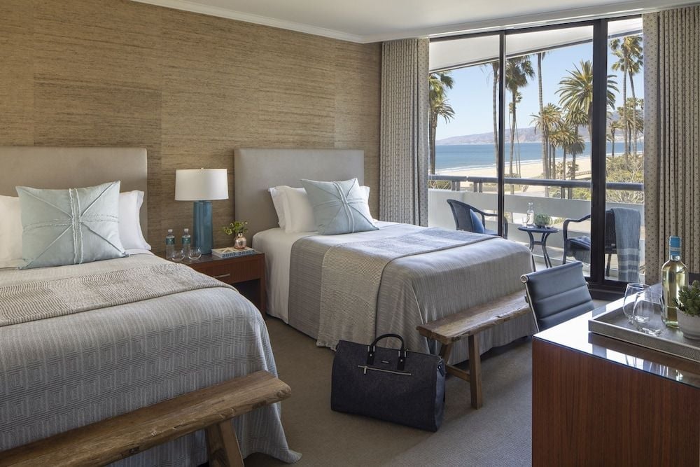 Twin beds draped in coastal linens frame panoramic Santa Monica views, where swaying palms and Pacific blue create an intimate retreat above the bustling beachfront below.