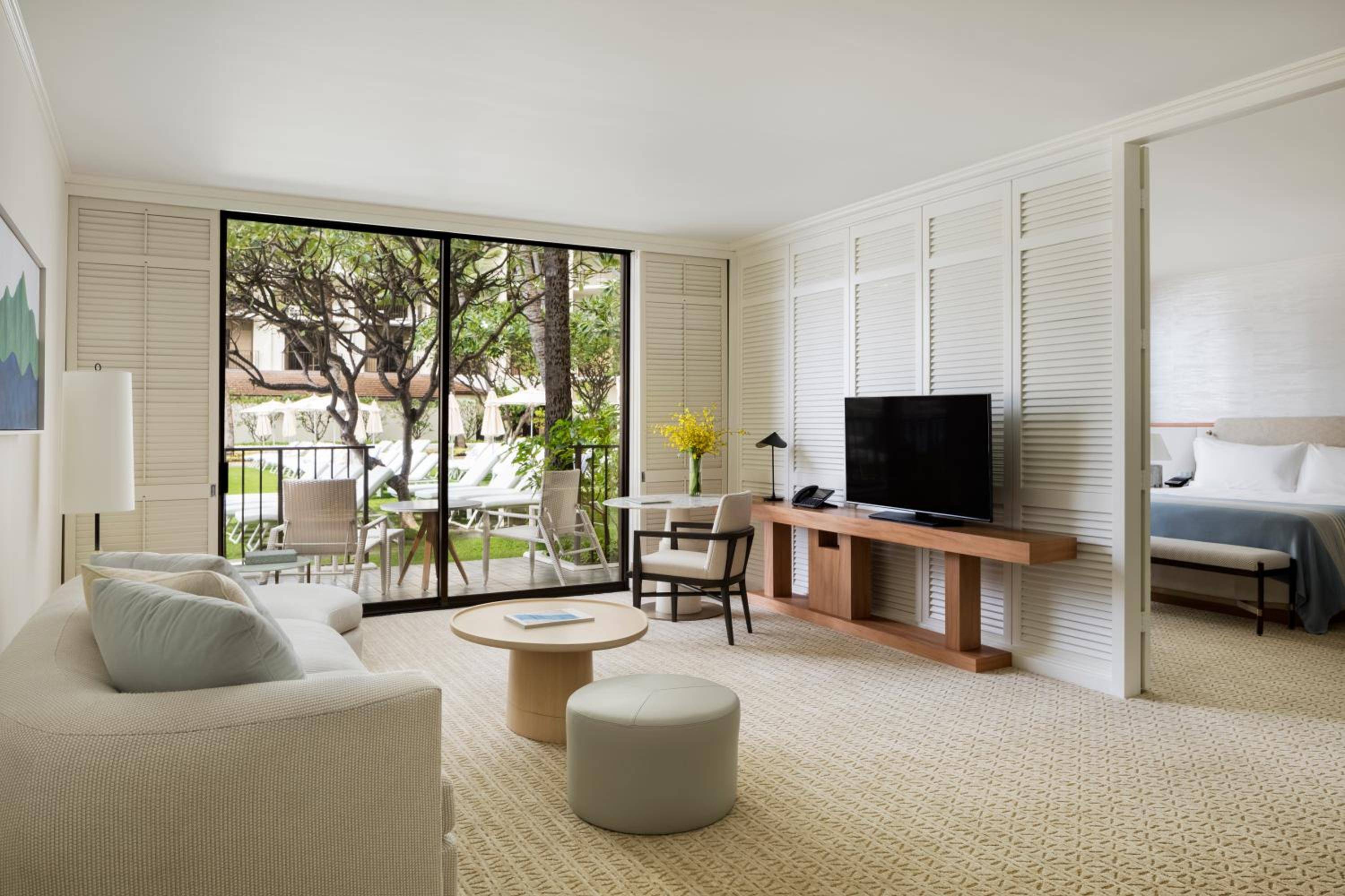 Natural light filters through bamboo blinds in this spacious suite, where warm wood furnishings and neutral linens create a serene Hawaiian retreat overlooking lush gardens.