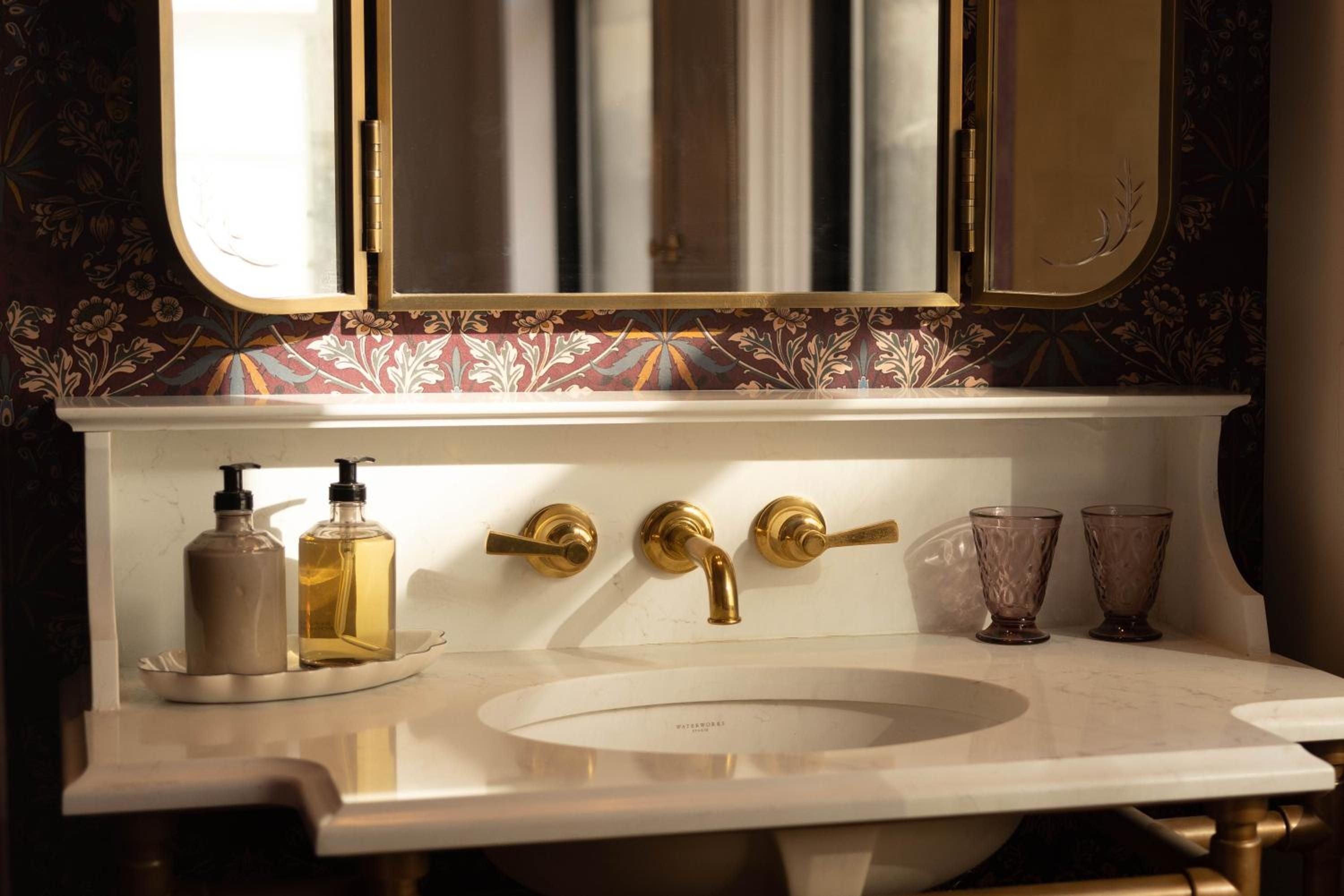 Brass fixtures gleam against William Morris-inspired wallpaper in this powder room where vintage amber bottles and cut-glass tumblers create an intimate apothecary atmosphere.