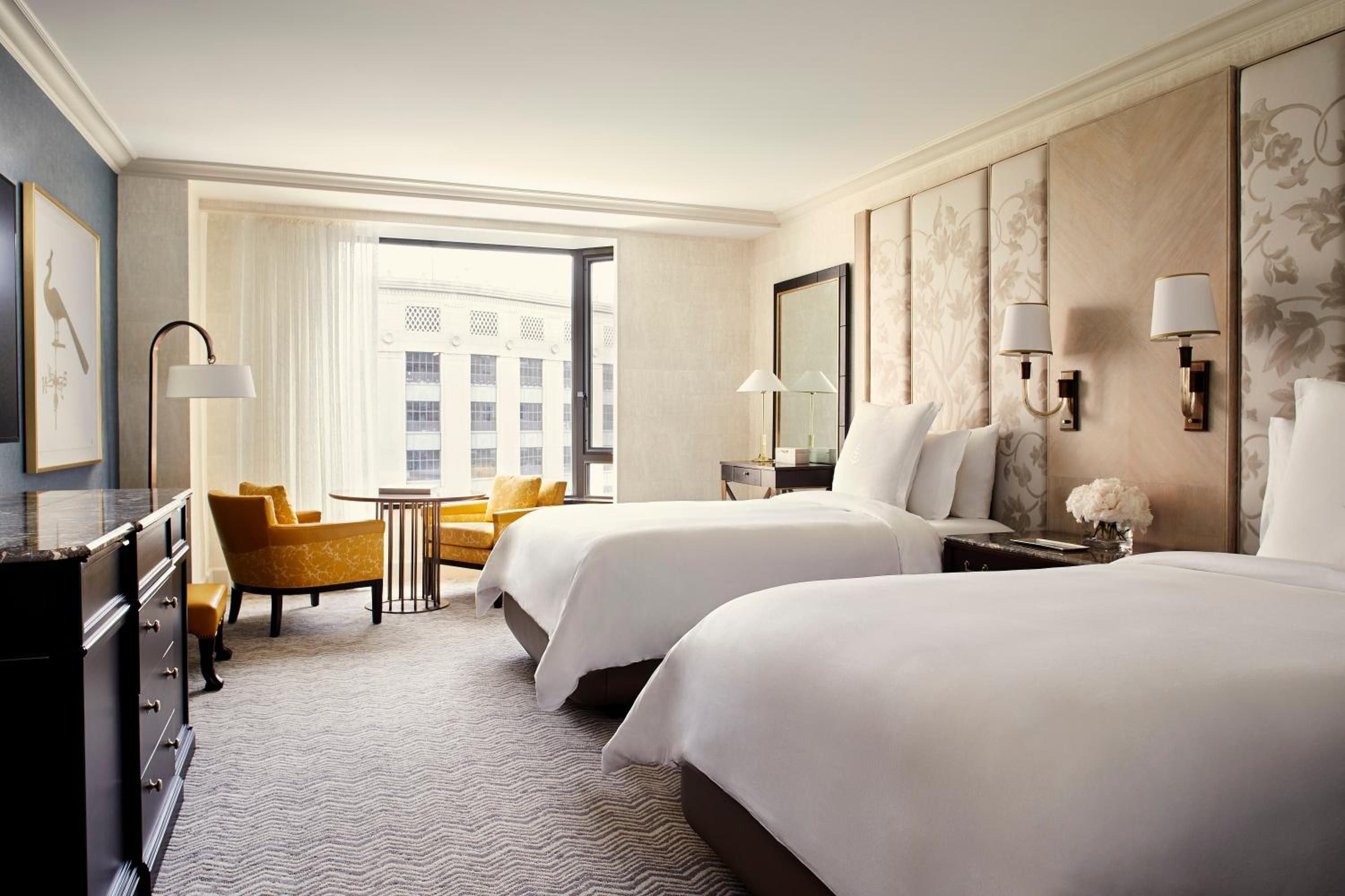 Crisp white linens meet warm taupe wallcoverings in this sophisticated twin bedroom, where golden velvet chairs catch afternoon light streaming through floor-to-ceiling windows.