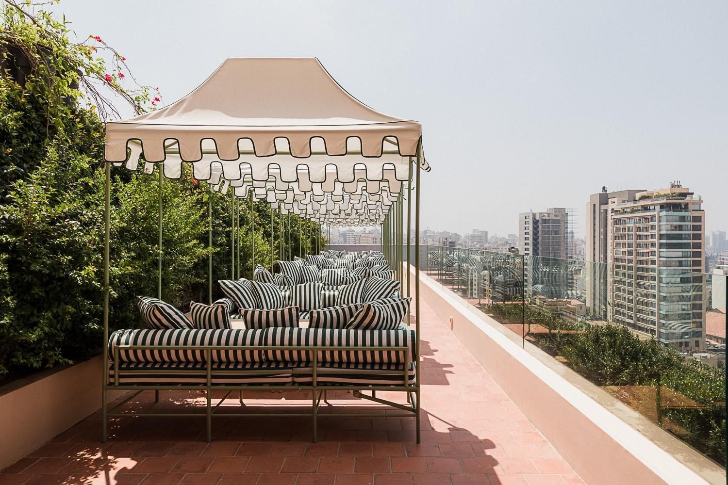 Black and white striped cushions create a cabana-style retreat on the rooftop terrace, where urban skyline views stretch beyond leafy green plantings and terracotta tiles.