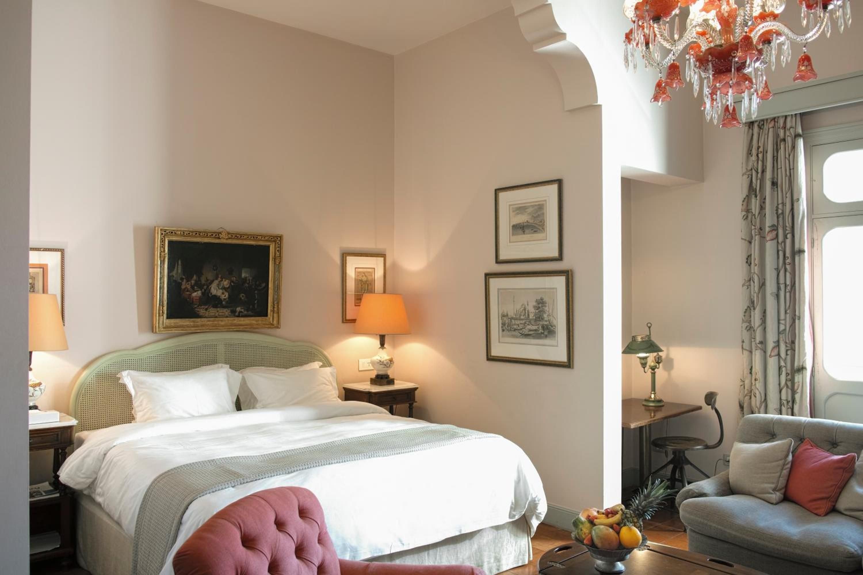 Warm lamplight illuminates classical artwork and plush linens in this elegantly appointed suite, where coral chandeliers cast romantic shadows across cream-colored walls.