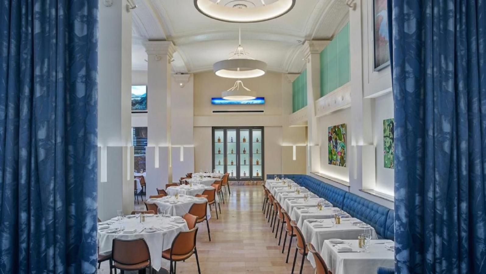 Crisp white linens and cognac leather chairs create an elegant canvas beneath soaring ceilings, while vibrant contemporary art adds unexpected pops of color to this sophisticated dining room.